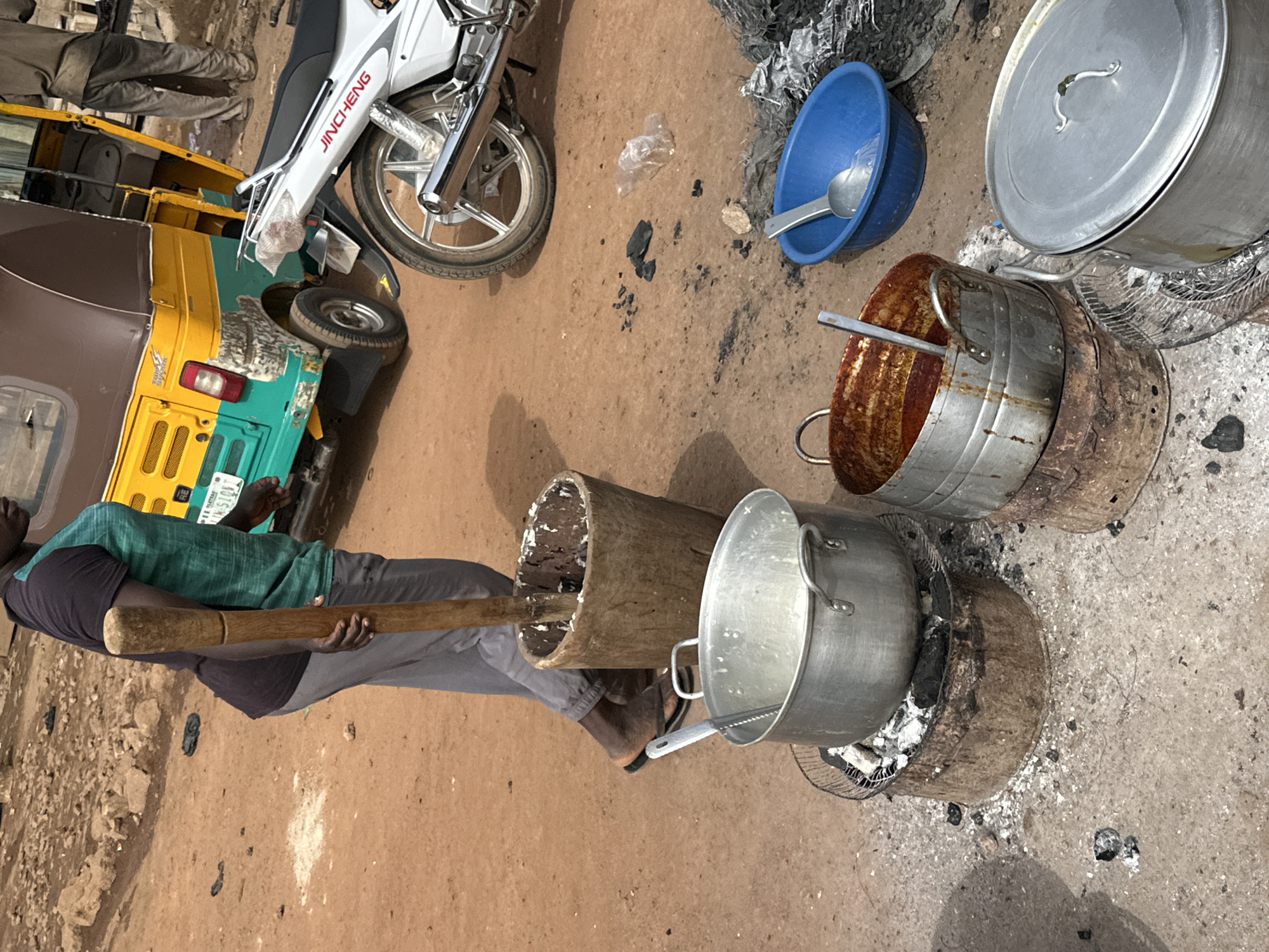 The image depicts an outdoor food preparation and cooking scene, likely in a rural or semi-urban area such as Gura Top, Nigeria. In the left foreground, a man, dressed in a green and black shirt and grey pants, is engaged in pounding something in a large wooden mortar with a long wooden pestle. His foot is visible near the base of the mortar.
To the right, several cooking vessels are arranged on the dirt ground. Two large aluminum pots are set over rudimentary stoves, which appear to be hollowed-out sections of wood acting as charcoal burners, with ash and embers visible. One of these pots contains a light-colored substance. Nearby, another large aluminum pot with a lid and a smaller, rusted metal pot are placed on the ground. A blue plastic bowl containing a spoon is also present.
In the background, a white motorcycle with "JINCHENG" written on its side is parked, alongside a yellow and green auto-rickshaw (tuk-tuk). The auto-rickshaw's license plate shows "RE" and partially visible numbers. The ground is a light brown earth, scattered with ash, charcoal pieces, and some debris, indicating an active outdoor workspace. The scene appears to be taking place during daylight hours under natural light.