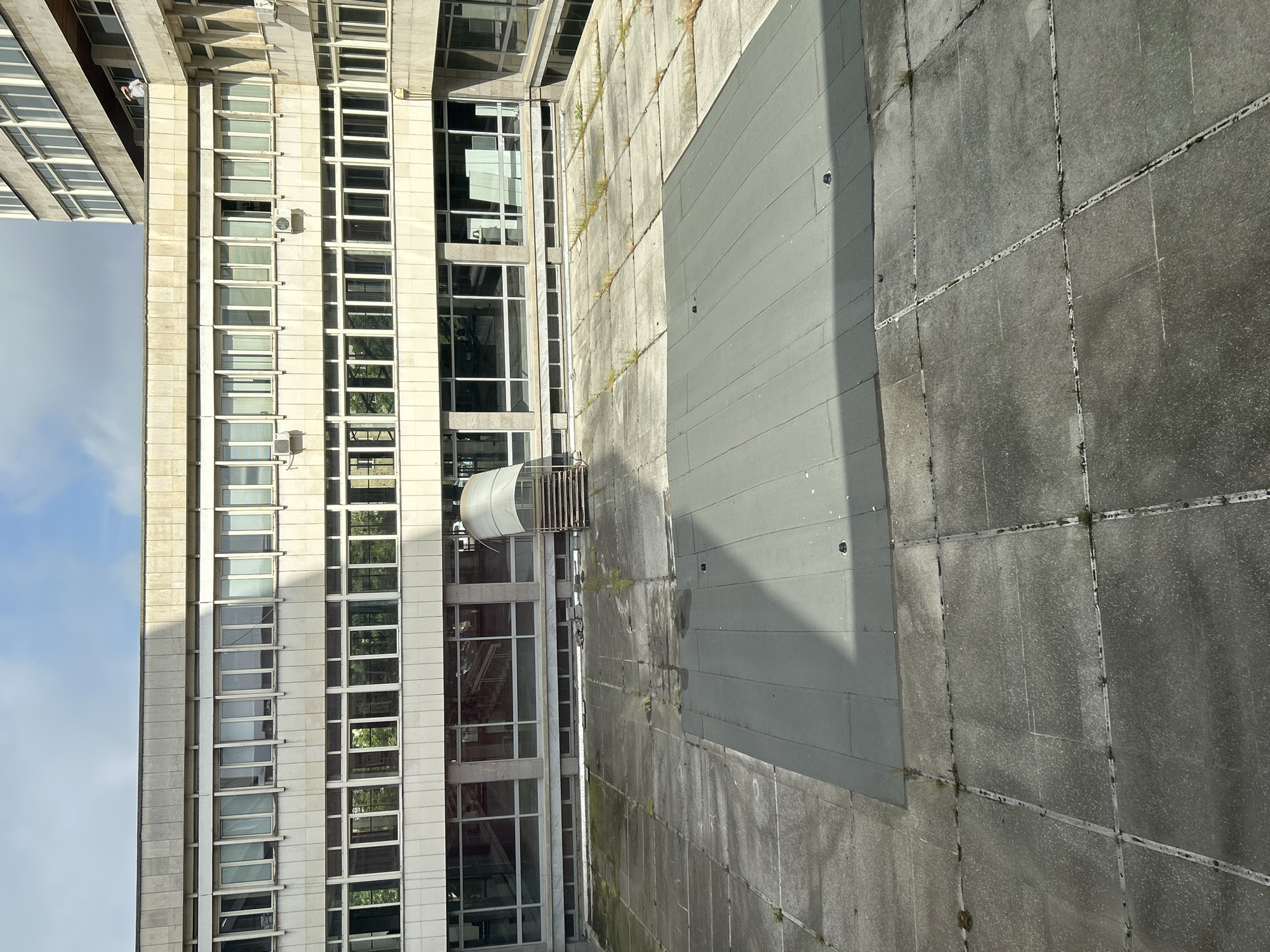 The image provides a high-angle perspective of an urban setting in Sofia, Bulgaria, showcasing a multi-story building alongside a paved outdoor area. The central subject is a modern, tall building on the left, featuring a facade composed of light-colored, possibly concrete or stone, vertical pillars and horizontal bands. Numerous large glass windows, some reflecting surrounding trees and structures, fill the spaces between these architectural elements. A few windows appear open, and several air conditioning units are visible on the exterior wall. In the upper part of the building, a small figure in a white top can be seen on a ledge, possibly engaged in maintenance or work.
To the right of the building lies a broad, flat paved area made of large, grey concrete slabs arranged in a grid pattern. The surface shows signs of weathering, with visible dirt and small patches of moss or weeds growing in the seams between the slabs. A prominent, dark grey rectangular platform or structure is situated on this paved surface, casting a shadow. Near the base of the building, a small, curved, translucent canopy with metal railings extends outwards, likely covering an entrance or a small outdoor space.
The sky, visible on the far left, is bright blue with scattered white clouds, indicating clear daylight. The strong, defined shadows cast by the building and the rectangular platform suggest a sunny day, likely mid-morning or afternoon. The overall scene conveys a functional, somewhat stark urban environment, with the only discernible human presence being the distant figure on the building. There is no visible text in the image.