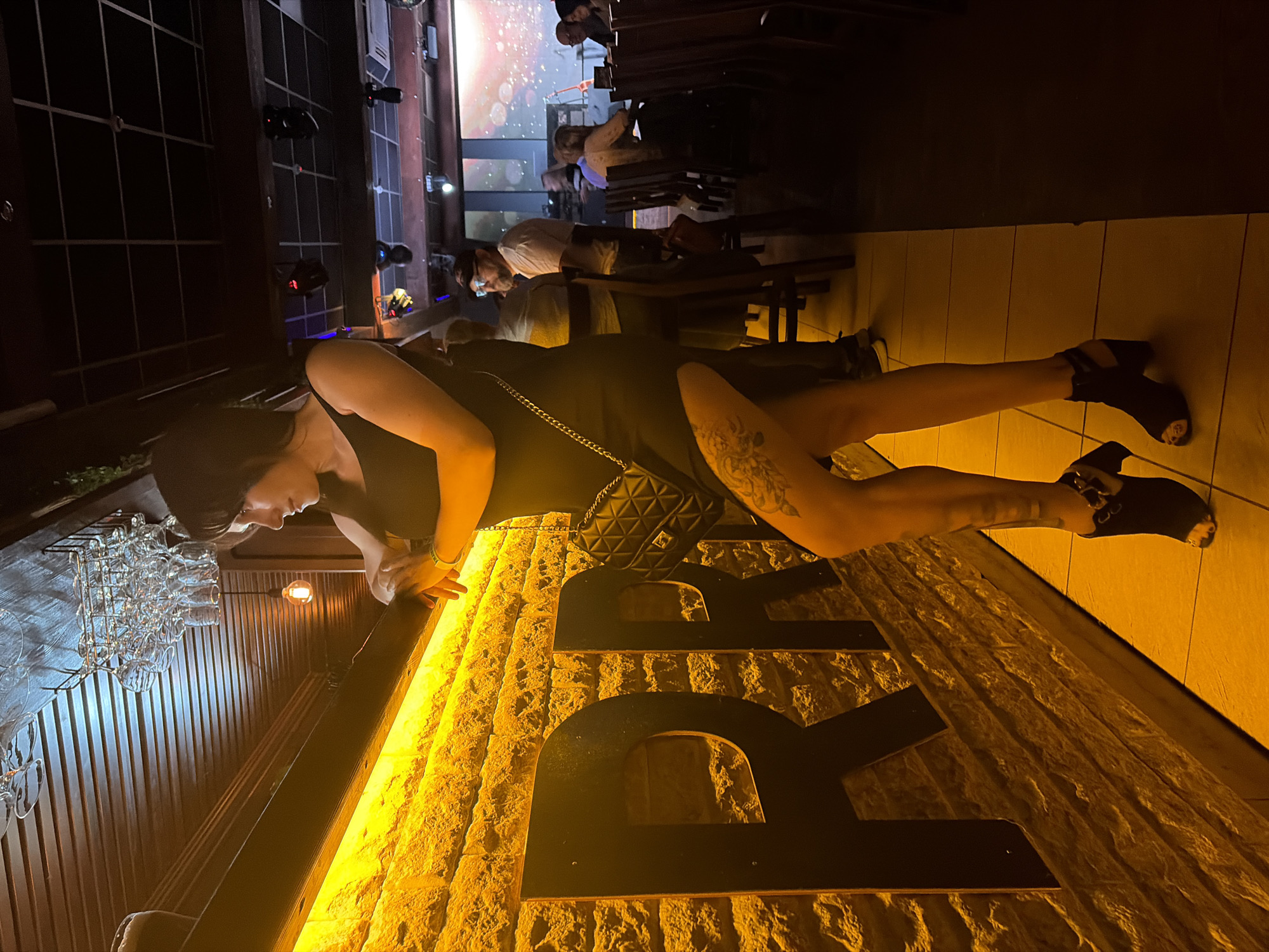 A woman in a black dress poses lying on a surface displaying the letters "BR". She wears black heeled shoes and a black purse with a chain strap. The background shows a bar or restaurant with tables and people. The location is consistent with a bar in the city of Chaikovsky, Russia based on other indicators.