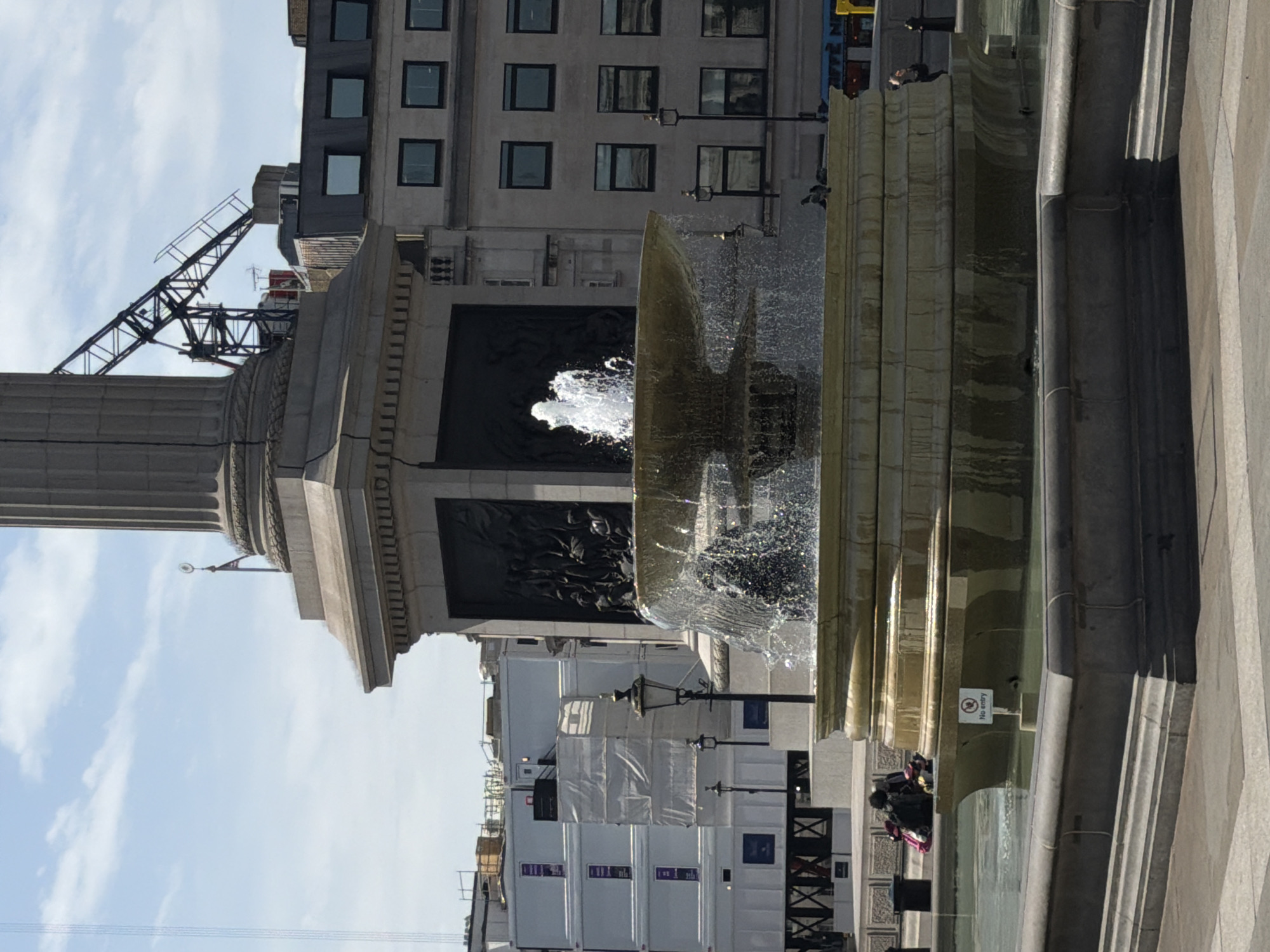The image shows a view of Trafalgar Square in London, United Kingdom. In the foreground is a fountain with water spraying upwards. To the left is Nelson's Column, a monument with a tall column structure. Behind the column and fountain, there are buildings with multiple windows. A construction crane is also visible near the top of one of the buildings. Several people are present near the fountain. The sky is visible with some clouds.