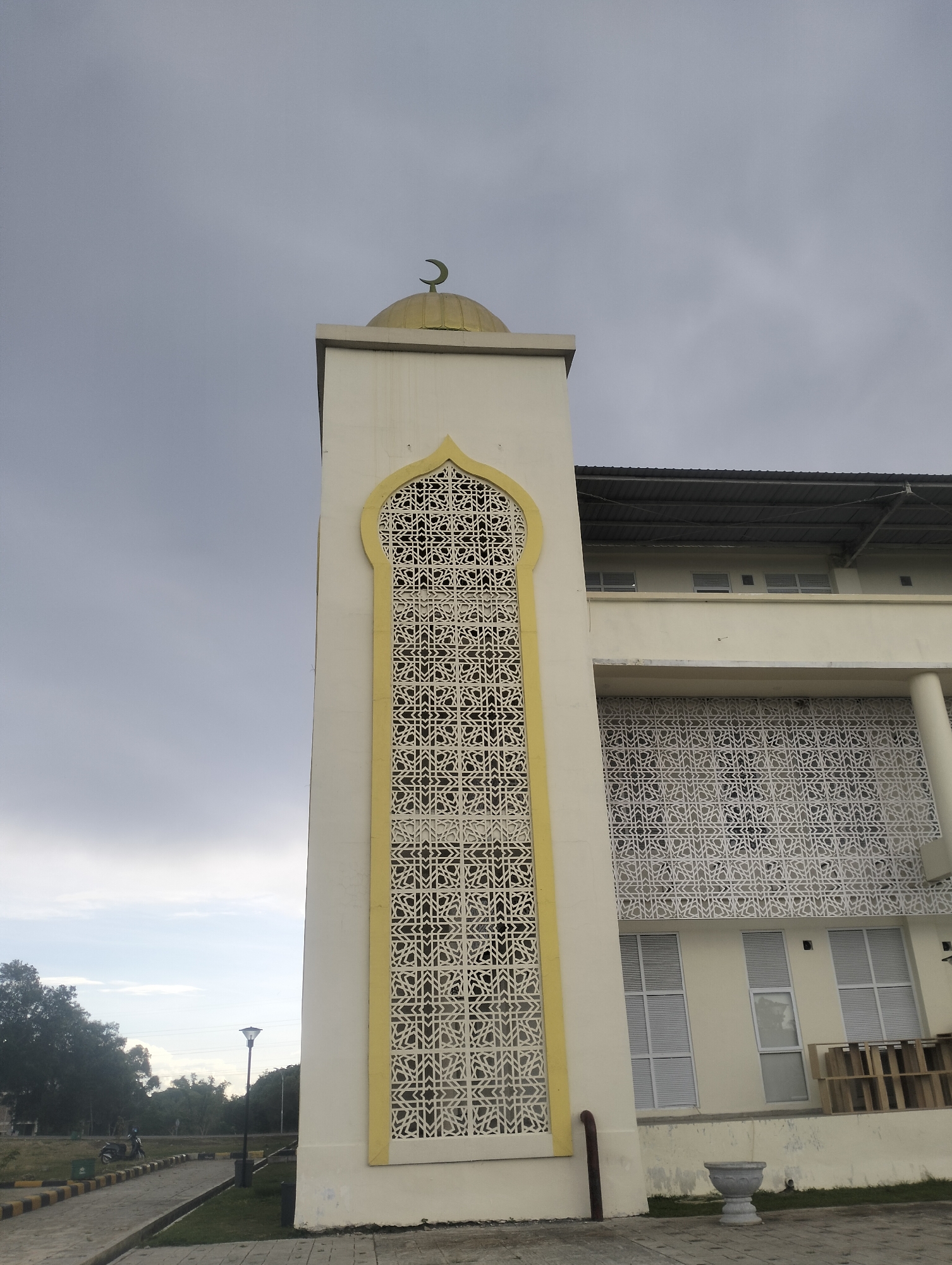 The image shows a close-up of a minaret of a mosque in Reuleut Timu, Indonesia. The minaret is tall and rectangular, painted white with yellow accents. It features an intricate, geometric pattern on its side, which appears to be a decorative screen. At the very top of the minaret, a golden crescent moon and star are visible against a cloudy sky. To the right of the minaret, the main building of the mosque is partially visible, also painted white and featuring similar decorative screens on its exterior. The building has multiple windows and a dark, sloped roof. In the foreground, to the left of the minaret, a paved walkway is visible, bordered by grass and a curb. Further in the background, trees and a street lamp can be seen. A motorcycle is parked on the side of the road. The overall impression is a modern mosque structure under an overcast sky.