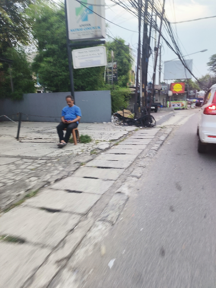 The image shows a man sitting on a wooden chair on a sidewalk. The sidewalk has a gray concrete surface with cracks and is located in front of a gray wall. There are a few green plants growing on the sidewalk. The man is wearing a light blue shirt and dark pants. He is looking down at something in his hands. There are power lines running along the top of the image. There is a white car traveling on the street to the right of the sidewalk. The car is partially visible and the driver's side is facing the viewer. The car is in the middle of the street. The street is paved with black asphalt and there is a gray concrete curb on the left. The location is a residential area with a small clinic on the left side of the sidewalk. The clinic sign states "Apotik Sumber Sehat," which translates to "Health Source Pharmacy." The image was taken at latitude -6.939 and longitude 107.737, which is located in the city of Cileunyi, Indonesia.