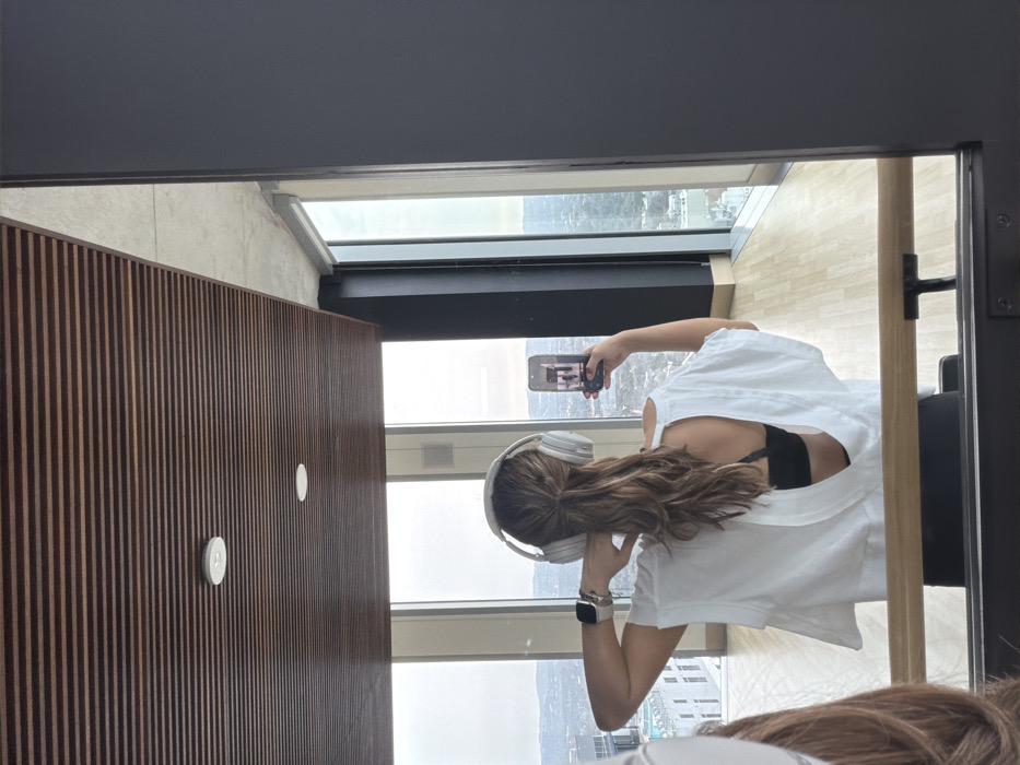 Inside a modern, high-rise apartment, a woman wearing white headphones and a white t-shirt is taking a picture with her phone of the cityscape outside the window. Her back is to the camera, and her hair is long and wavy. A wood-paneled wall is on the left, and a doorway with a black frame is on the right. The sky outside the window is overcast.
