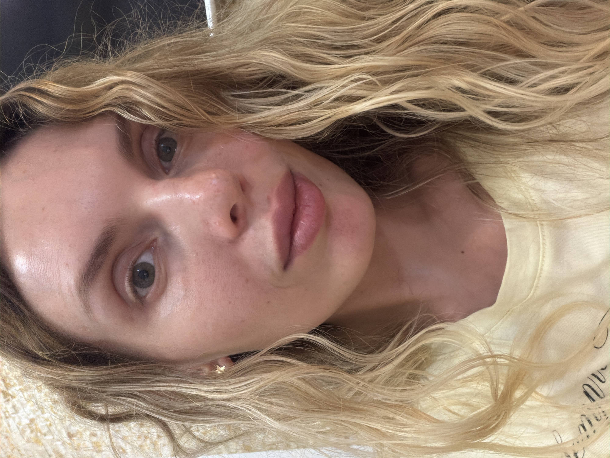 A close-up, angled headshot of a fair-skinned woman with shoulder-length, wavy blonde hair is presented. Her facial features include light gray-green eyes, a small nose, and full lips. She has a small gold earring on her left ear. She's wearing a light yellow shirt, and the texture of the background underneath her hair suggests a fabric surface, possibly a blanket. The scene is not explicitly identifiable with Budva, Montenegro, or any other specific location. There are no discernible activities, nor interactions between people. The lighting appears natural and bright, illuminating the woman's face and hair.