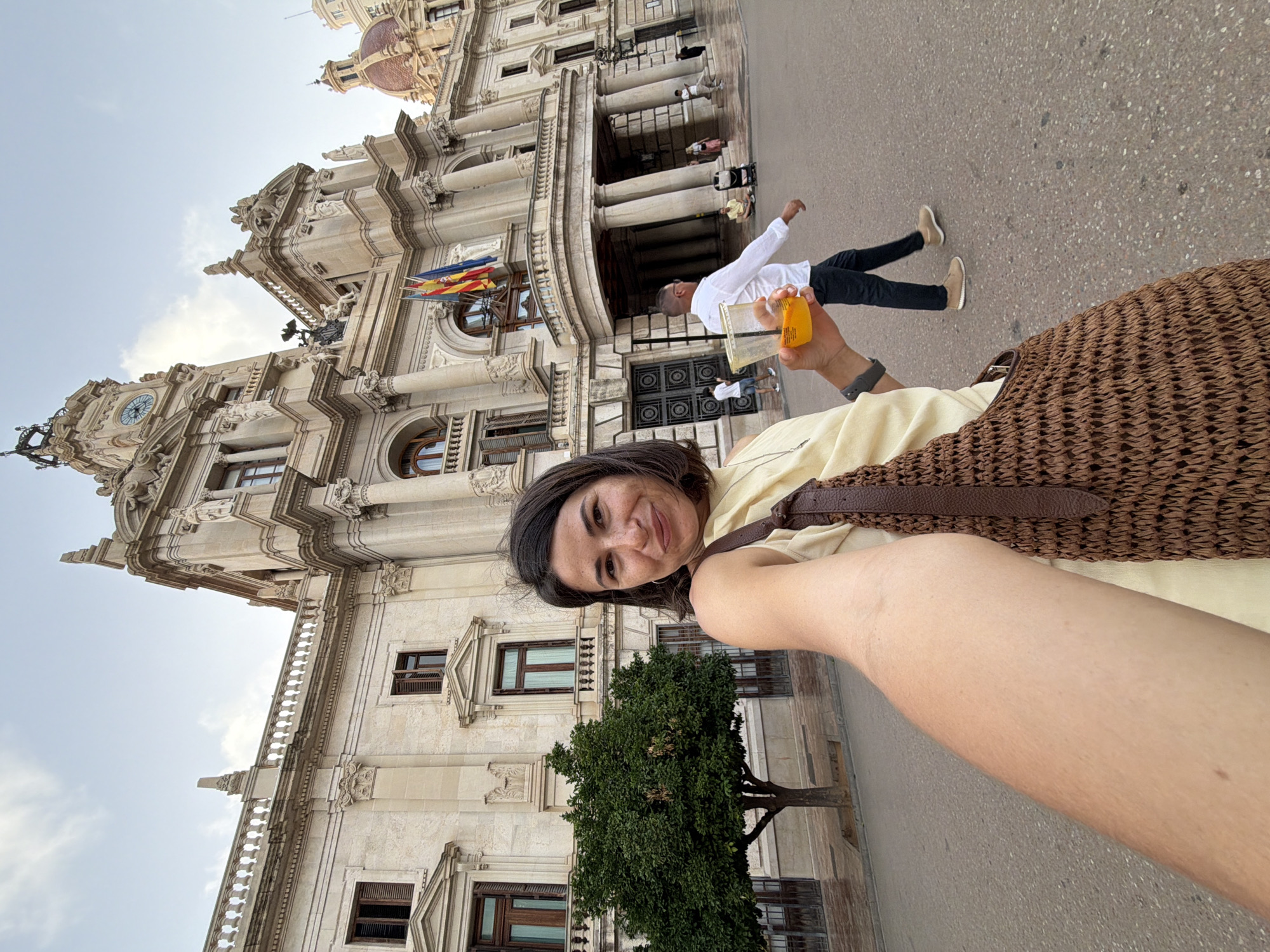 In an urban outdoor setting, a woman with dark hair is taking a selfie in front of a large, ornate building. She is wearing a light yellow top and carrying a brown woven bag over her shoulder. In her right hand, she holds a clear plastic cup filled with an orange beverage. Behind her, the building features classical architecture with columns, arches, and flags. A small, bushy green tree is to her left. To her right and further in the background, a man in a white shirt and dark pants walks away from the camera. Other people and a stroller are visible in the distance, suggesting a public space. The ground appears wet, possibly from recent rain. The sky is overcast.
