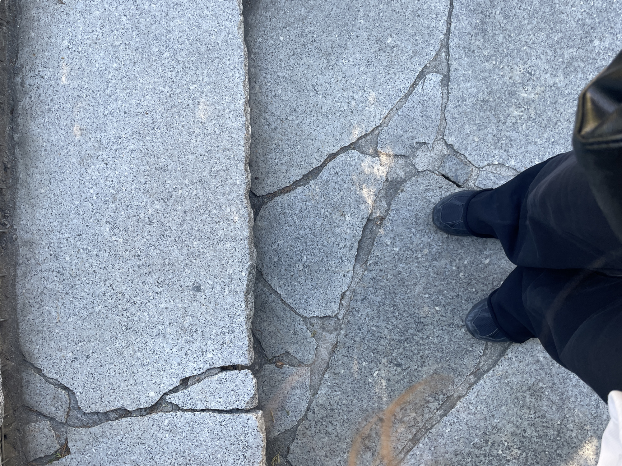The image depicts a section of damaged granite pavement in an unknown urban location. The pavement is cracked in multiple places, with visible lines indicating the breaks. A person wearing dark pants and dark shoes is standing on the cracked pavement. The left side of the image shows a larger, uncracked granite block adjacent to the broken section. There is some debris visible within the cracks of the pavement. The lighting suggests it is daytime.