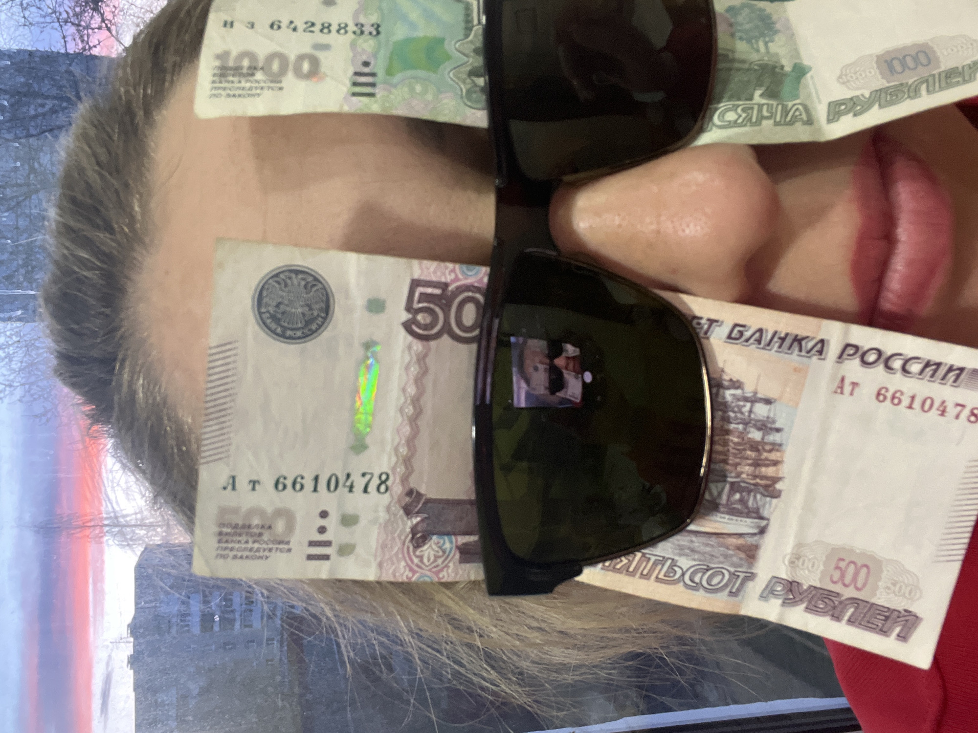 The image presents a close-up, portrait-oriented shot of a person with Russian ruble banknotes strategically placed on their face, partially obscuring it. The person is wearing dark sunglasses, with a 1000 ruble banknote (marked "1000 РУБЛЕЙ", serial number "ИЗ 6428833") draped across the forehead and a 500 ruble banknote (marked "500 ПЯТЬСОТ РУБЛЕЙ", serial number "АТ 6610478") covering the cheek and nose. The 500 ruble note also bears the warning "ПОДДЕЛКА БАНКА РОССИИ ПРЕСЛЕДУЕТСЯ ПО ЗАКОНУ" (Counterfeiting by the Bank of Russia is prosecuted by law).
The lower part of the face, including lips with pink lipstick, is visible below the sunglasses and banknotes. Light-colored hair frames the face. The dark lenses of the sunglasses show blurry reflections, possibly of a room interior or another individual.
The background, seen through a window, displays an outdoor scene with a sky featuring soft pink, purple, and blue tones, indicative of either sunrise or sunset. Bare trees or branches are silhouetted against the sky, suggesting a cold season like winter. The scene, located in Chaikovsky, Russia, uses Russian currency as a prominent element, conveying a playful or symbolic message related to money.