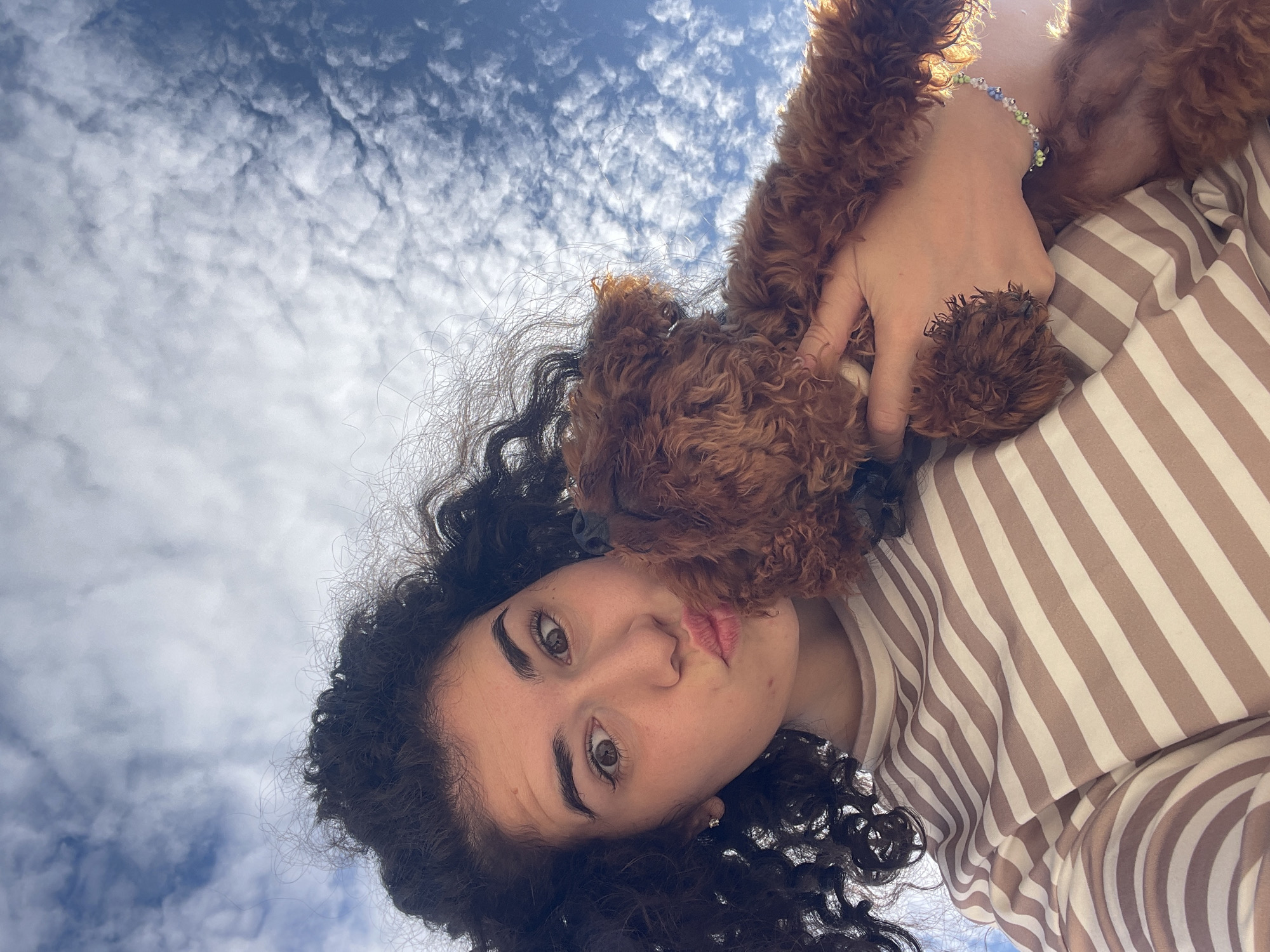 A low-angle upward-facing image captures a young female and a small dog against a backdrop of a blue sky with scattered white clouds, located in Koprinka, Bulgaria. The female, with dark curly hair, brown eyes, and fair skin, is positioned on the left side of the frame, looking directly at the camera with wide eyes and a puckered lip expression. She wears a horizontal striped long-sleeved shirt in alternating beige and white bands. A small, clear stud earring is visible in her left ear. A small, fluffy, reddish-brown dog is held by her in the right half of the frame. The dog's head is near the female's face, with its nose close to her lips. The female's left hand supports the dog from below. A multicolored beaded bracelet, featuring light blue, green, and white beads, is visible on her right wrist. The upper portion of the image displays a clear blue sky interspersed with numerous small, textured white clouds.