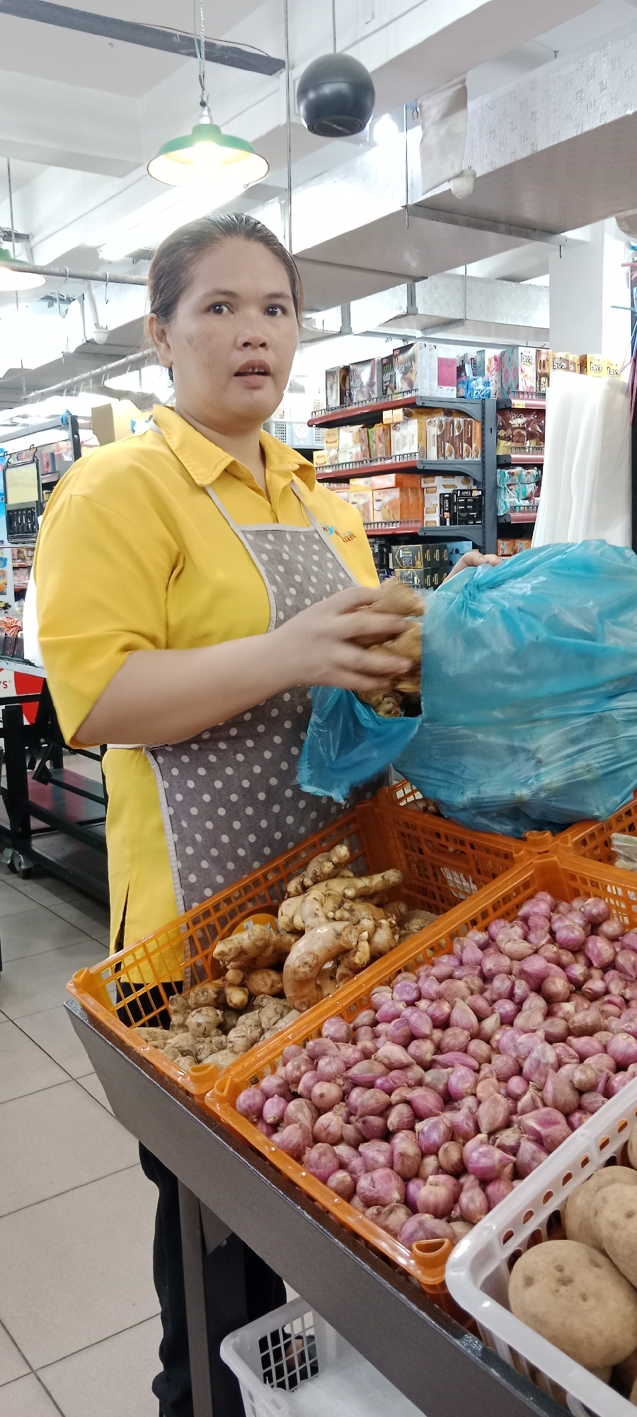 A woman wearing a yellow shirt and a grey polka dot apron is standing in a supermarket in Tanjung Pinang, Indonesia. She is holding a bunch of ginger root in her hands, which are partially inside a blue plastic bag. In front of her are two orange plastic crates filled with produce. The crate on the left contains more ginger, while the crate on the right is filled with shallots. To the right of the shallots, there is a white basket with potatoes. In the background, shelves are stocked with various food products. The scene appears to be within the produce section of a grocery store.
