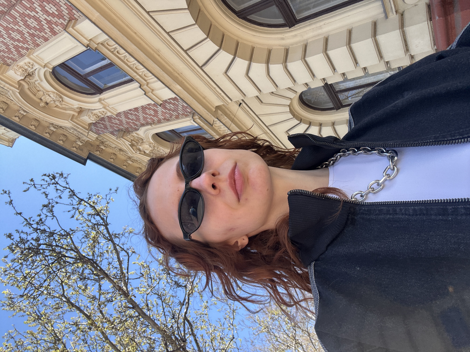 A low-angle shot depicts a young woman with reddish-brown curly hair, sunglasses, and a black jacket, posing outdoors in Novi Sad, Serbia. She is wearing a white shirt and a silver chain. The background includes an ornate building with windows, beige and red brick details, and the blue sky is visible above the tree branches. The photo is taken from below, looking up at the subject.