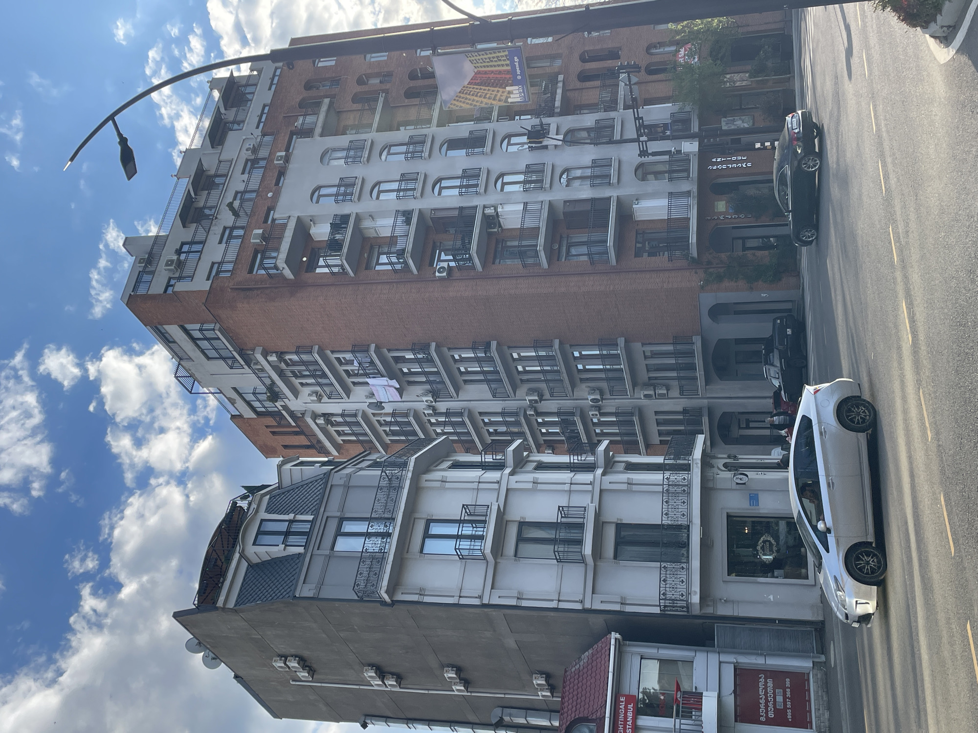 An outdoor, slightly rotated photograph captures a vibrant urban street scene in Tbilisi, Georgia, under a bright, partly cloudy sky. The main focus is a street lined with multi-story buildings on the left and a paved road with vehicles on the right.
The buildings vary in architectural style, featuring a mix of modern and traditional elements. A prominent central building displays a reddish-brown brick facade on its lower levels, transitioning to a light grey or white upper section with numerous windows, many adorned with black metal grilles or small balconies. Many air conditioning units are visible on its exterior walls. Above this building, a billboard shows a building design and the text "apage.ge" and "WE CARE". Another building closer to the foreground has a similar light-colored facade with more ornate black metal balconies and large ground-level windows. A section of a dark grey, possibly tiled, pitched roof is visible on this building.
On the right side, the road appears to be a two-lane street with white and yellow markings. A white hatchback, possibly a Toyota Prius, is prominent on the road closer to the viewer, while a dark-colored sedan is parked further down the street near another building. Several indistinct figures can be seen near a building entrance in the mid-ground.
Visible text on shop signs confirms the location. On a red sign, "NIGHTINGALE ISTANBUL" is partially legible along with Georgian text which reads "ფანჯრები და კარებები" (Windows and Doors) and a phone number "+995 597 386 399". Another sign displays "არქიმედეს" (Archimedes) in Georgian. The time of day is sunny daytime, indicated by the bright sky and clear lighting.