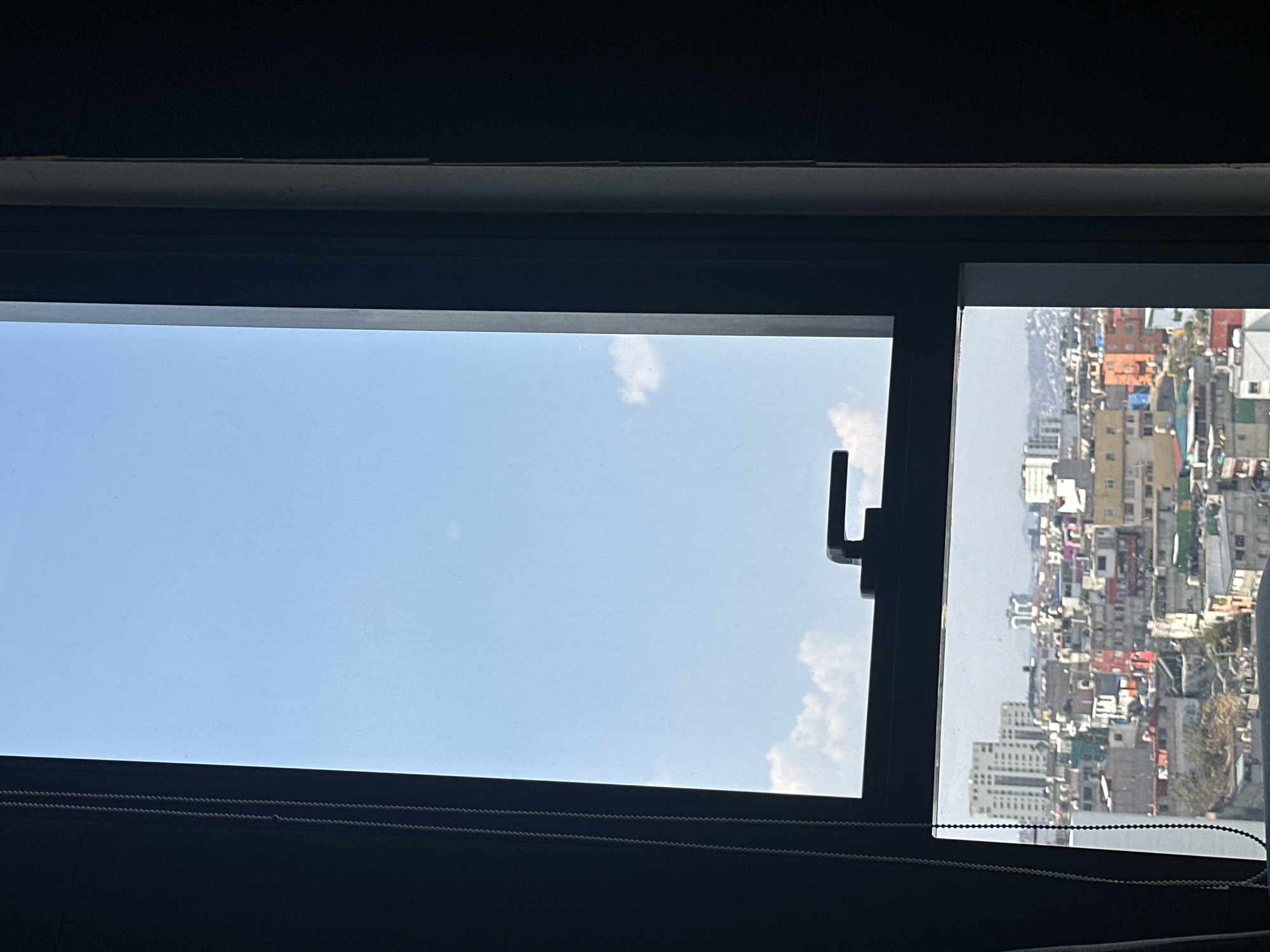 A window view shows a clear blue sky and cityscape in Huixquilucan de Degollado, Mexico. The sky is visible through the larger pane, while the city buildings are seen through a smaller pane to the right. A dark window frame surrounds the view. A window handle is visible on the frame between the panes. Strings of a window blind can be seen in the lower section of the view.