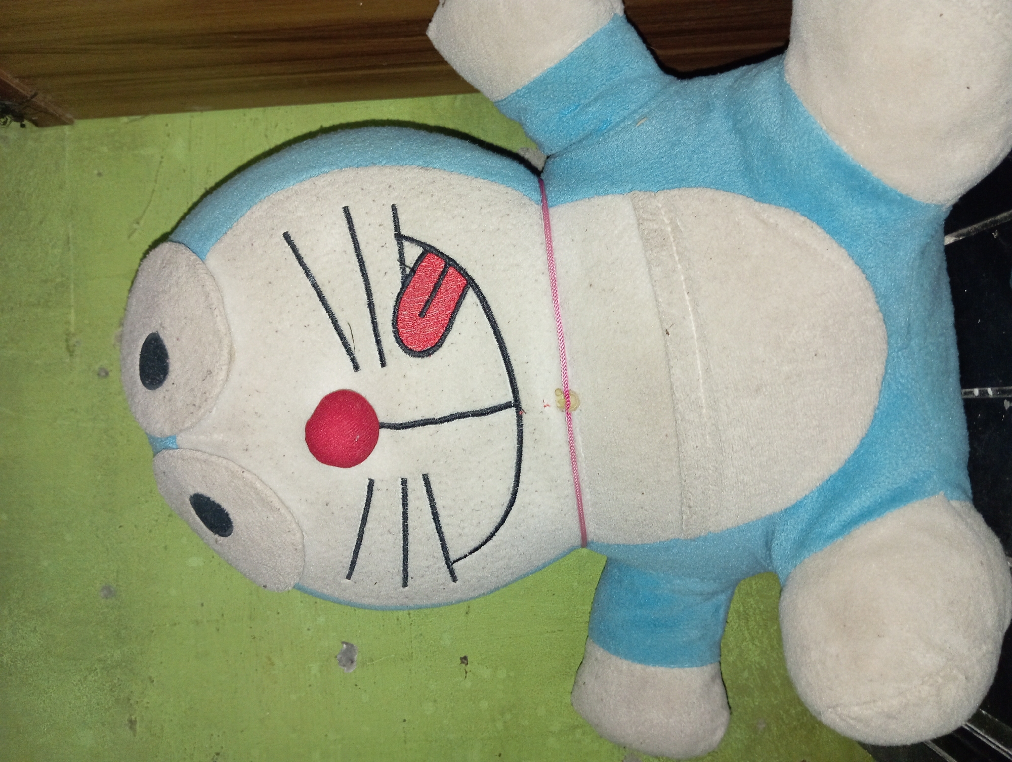 A stuffed toy resembling the cartoon character Doraemon is positioned against a green, textured wall. The toy is predominantly blue and white, with black embroidered facial features, including eyes, whiskers, and a smiling mouth with a red tongue sticking out. A thin pink string is wrapped around the toy's neck. The background wall shows signs of wear, with several small gray spots. To the right of the toy, a portion of a wooden surface, possibly a door or furniture, is visible. The setting is indoors, with the lighting suggesting artificial illumination. The scene appears to be a domestic environment within the city of Rejosari, Indonesia.