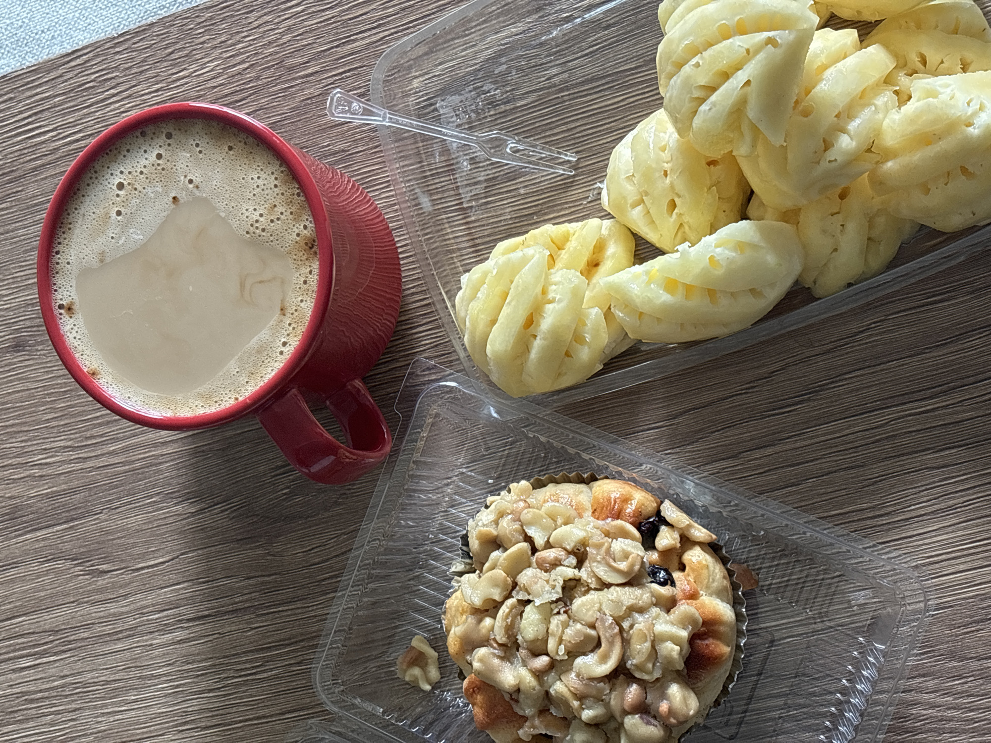 A red mug of coffee sits on a wood-grained surface. Adjacent to the mug is a clear plastic container holding cut pineapple. Below these items is another clear plastic container with a cashew-covered pastry. The scene is in Choeng Thale, Thailand.
