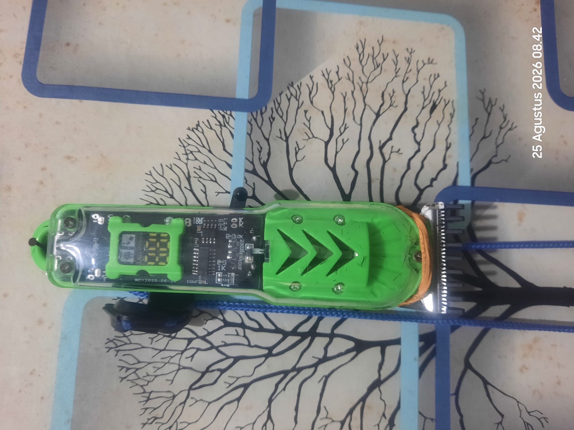 A green electric trimmer with a digital display is positioned vertically against a wallpaper depicting stylized trees. The trimmer has a transparent green casing, revealing electronic components, including a circuit board and a small screen showing yellow digital numbers. A blue cord is attached to the trimmer's top. The wallpaper features a light background with black tree branches and leaves. The bottom right corner of the image displays a timestamp: "25 Agustus 2026 08.42". The location is identified as Sukokerto, Indonesia.