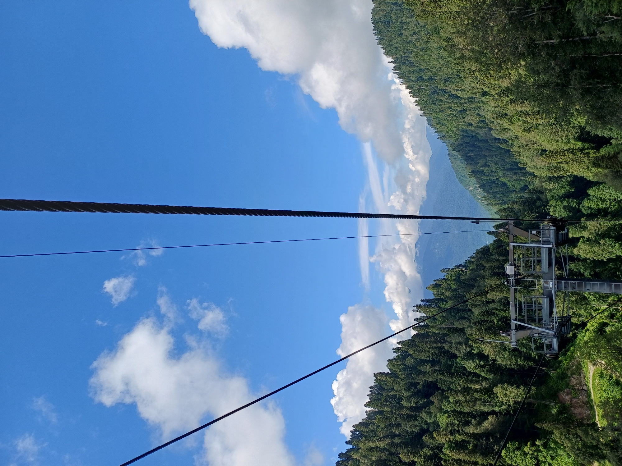 This is a photograph taken from a cable car ascending a mountain, offering a view of the forested slopes of Estosadok, Russia. The sky is a vibrant blue with scattered white cumulus clouds. In the foreground, thick black cables of the cable car extend upwards and diagonally across the frame. A segment of the cable car's support structure, a metal framework with wheels, is visible on the left side of the image. The mountain slopes are densely covered with a variety of evergreen trees, creating a textured green landscape that recedes into the distance. In the far background, layers of hazy blue mountains are visible under the bright sky. The scene depicts a scenic natural environment, likely a tourist destination for hiking or recreation.