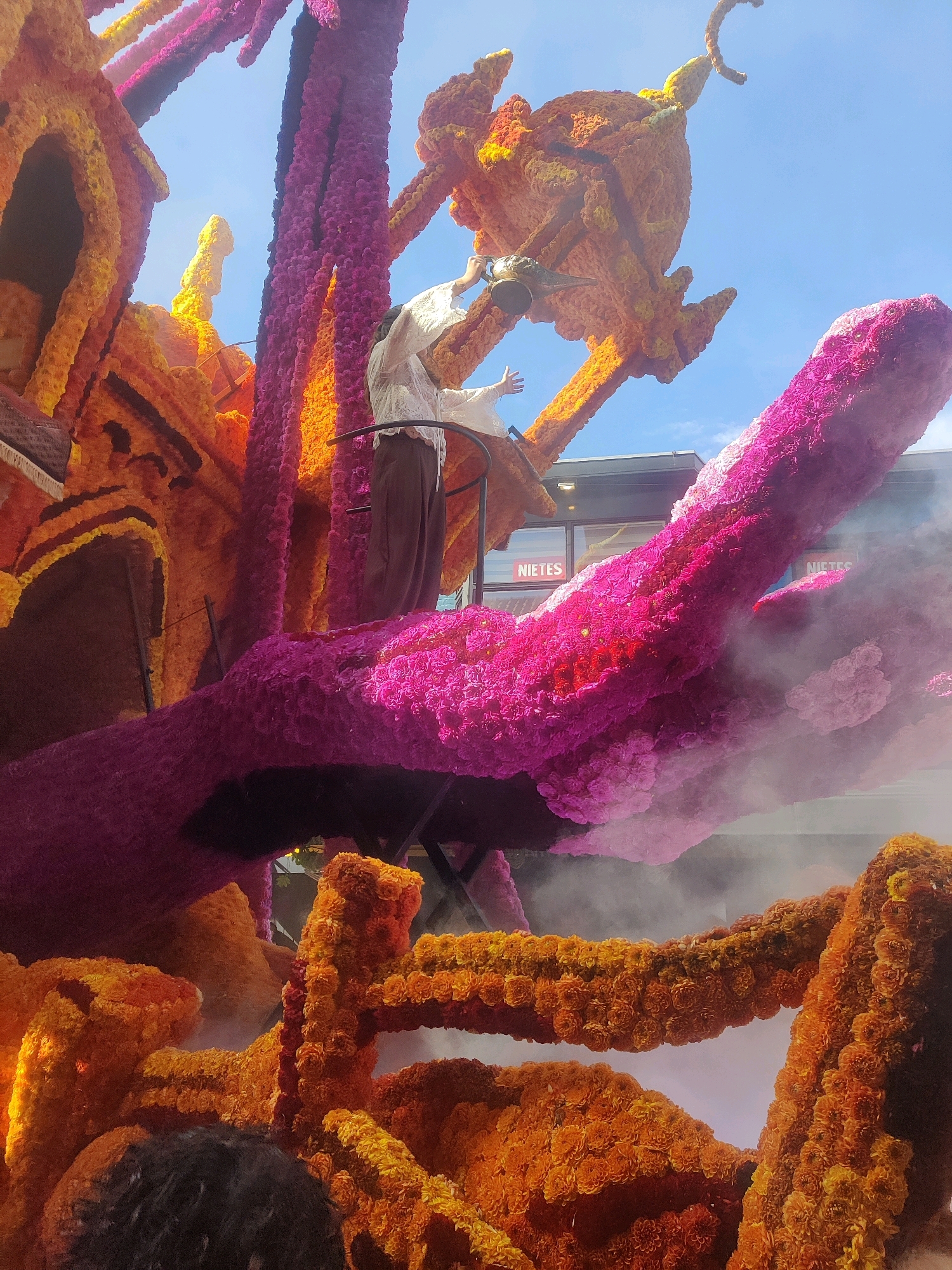 The image captures a vibrant floral float decorated with orange, yellow, and pink flowers, likely during the Zundert Flower Parade in Zundert, Netherlands. A person wearing a white, lacy top and brown pants is standing on the float, holding a golden lamp, possibly mimicking a genie from a story. The float features elaborate structures that resemble arches and perhaps a mythical creature, all intricately covered in thousands of flowers. Wisps of smoke or mist add a dramatic effect to the scene. In the background, a building with windows displaying the word "NIETES" is visible, along with a bright blue sky. The scale of the float and the detailed floral arrangements suggest a significant event.