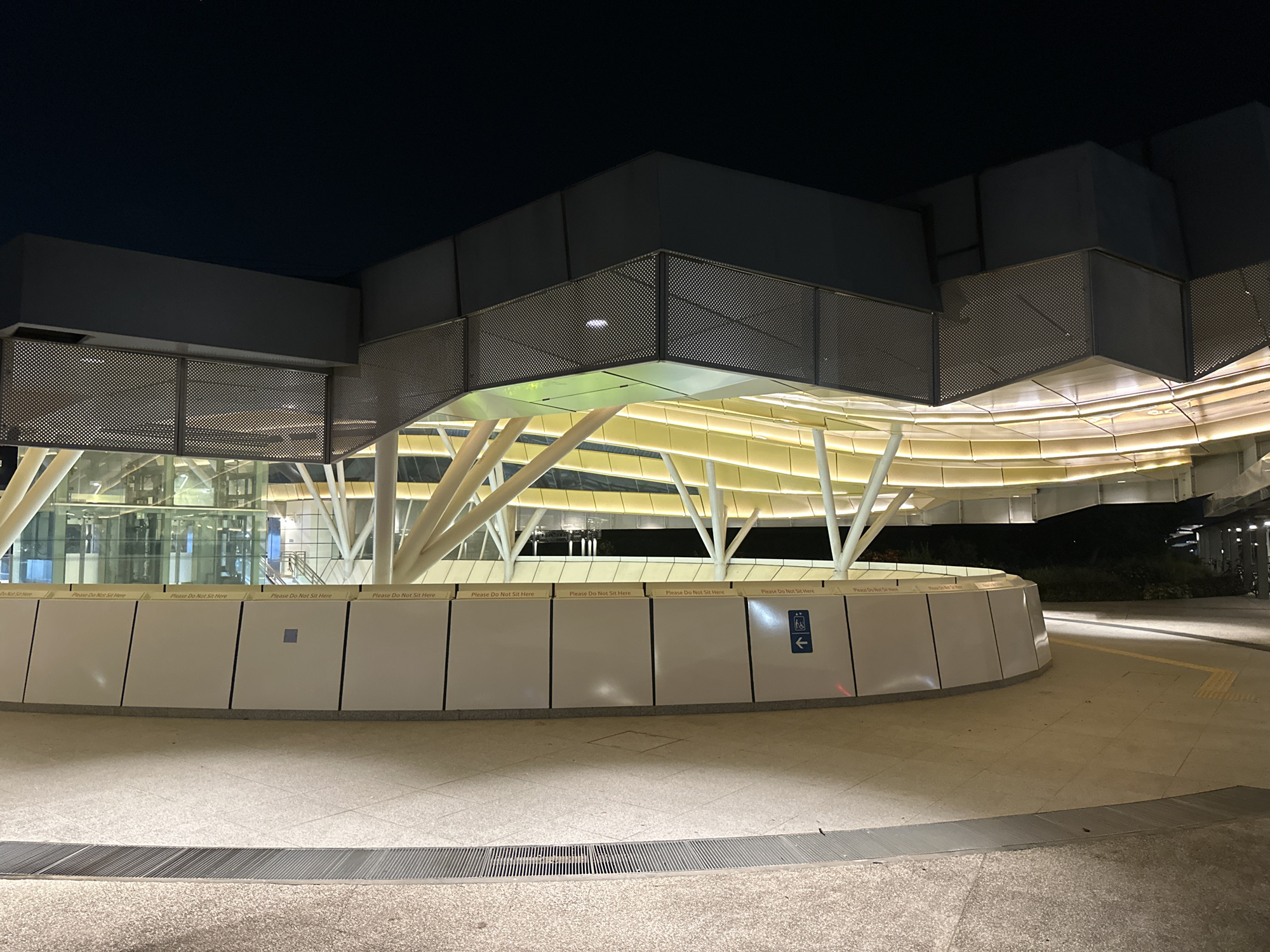 The image depicts the exterior of a modern, multi-level building at night in Singapore. The structure features a large, curved facade clad in white panels, with prominent, white, angled support columns. The upper levels are characterized by a perforated metal railing and dark, angular architectural elements. Yellowish-white lights illuminate the underside of the upper structure, creating a warm glow. A blue directional sign indicating accessibility is visible on the white facade. The ground level is paved with speckled concrete, with a metal grate running along the foreground. The sky is dark, indicating nighttime. The overall scene is clean and contemporary, suggesting a public transportation hub or a modern commercial building.