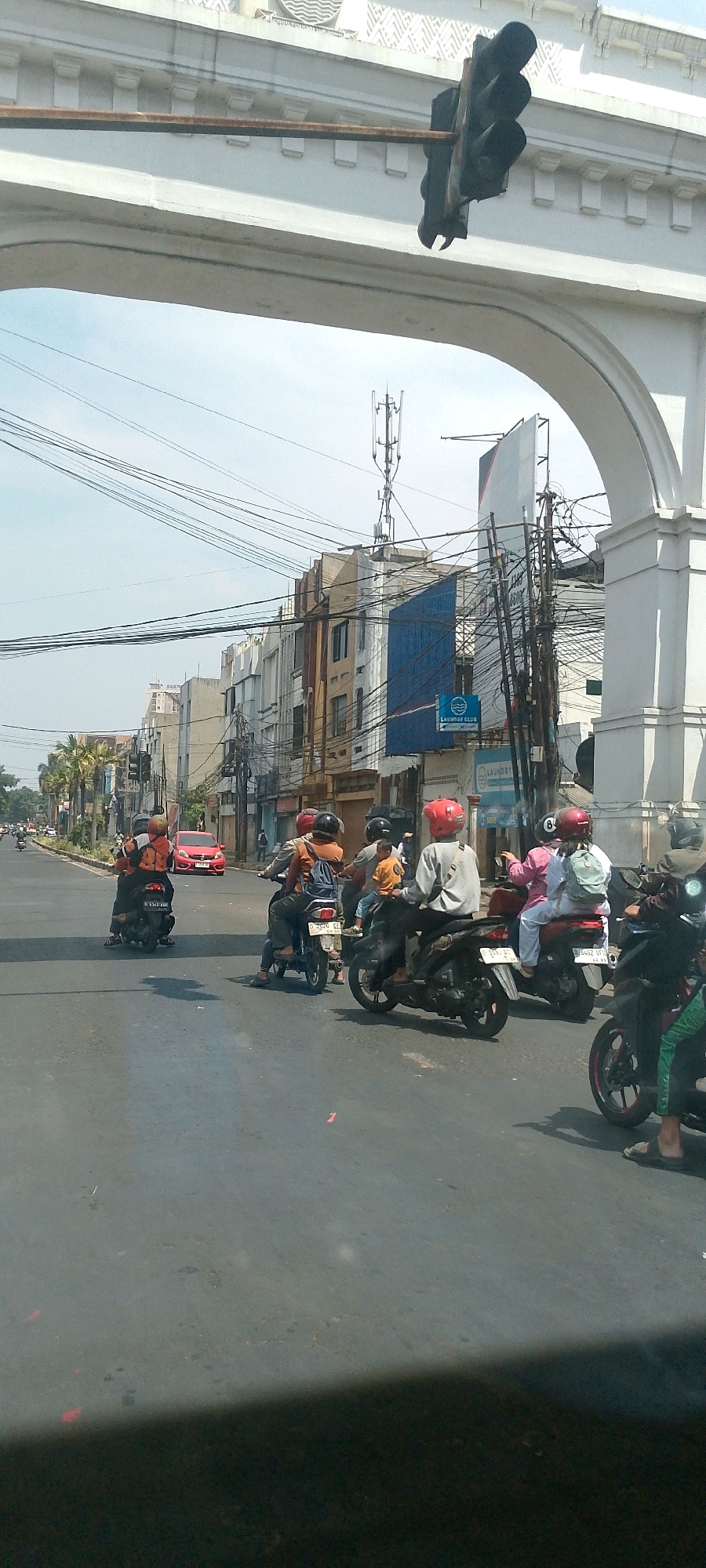 This image captures a street scene in Bandung City, Indonesia, likely on a sunny day with a bright sky and scattered clouds. The perspective is from within a vehicle looking forward and slightly upwards. Dominating the upper portion of the frame is a large, ornate white archway, possibly part of a building or a bridge, with intricate decorative elements. A rusted metal pole extends horizontally from beneath the arch, supporting a dark traffic light assembly.
Below the arch, a multi-lane road is visible, filled with various vehicles and pedestrians. The road surface appears to be asphalt, with some scattered debris and tire marks. Several motorcycles are prominent, with riders wearing helmets and casual clothing. Some motorcycles are carrying multiple passengers. In the middle ground, a red car is also present.
The background features a dense urban environment with multi-story buildings lining the street. These buildings vary in architectural style and color, with some appearing older and others more modern. Numerous electrical wires and communication antennas are visible strung across the cityscape, indicating a well-connected urban area. Palm trees and other vegetation are interspersed among the buildings, suggesting a tropical or subtropical climate. The overall impression is a busy, active street in a developed city.