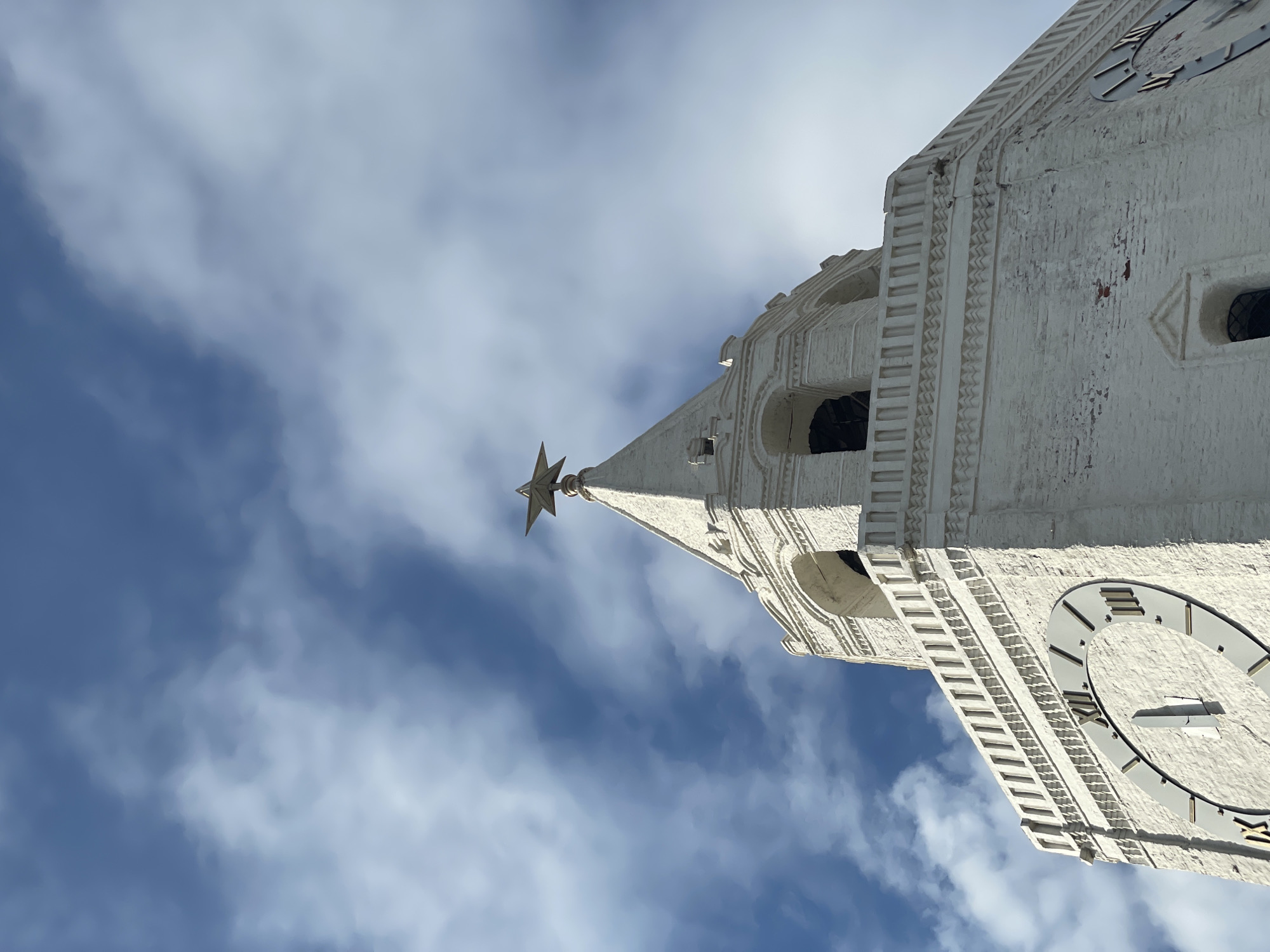 The image presents a striking upward view of a grand, white architectural tower against a bright, partially cloudy blue sky. The tower, likely made of stone or painted brick, showcases intricate architectural details, including decorative molding, textured surfaces, and arched openings.
A large clock face with dark Roman numerals is a prominent feature on one side of the tower. Visible numerals include XII, I, II, X, XI, and V, suggesting a time around noon or midnight, though the exact time is not clearly discernible from the hands.
Crowning the slender spire at the very top is a distinctive five-pointed star finial, which appears golden or reddish under the sunlight. This star, along with the specific architectural style and clock, strongly suggests the tower is the Spasskaya Tower of the Moscow Kremlin.
The sky is a vibrant blue, adorned with soft, scattered white clouds, indicating clear, daytime weather. The low-angle perspective emphasizes the imposing height and historical grandeur of the structure. No human activity or specific events are depicted, focusing solely on the architectural subject. The scene evokes a sense of historical significance and architectural beauty.