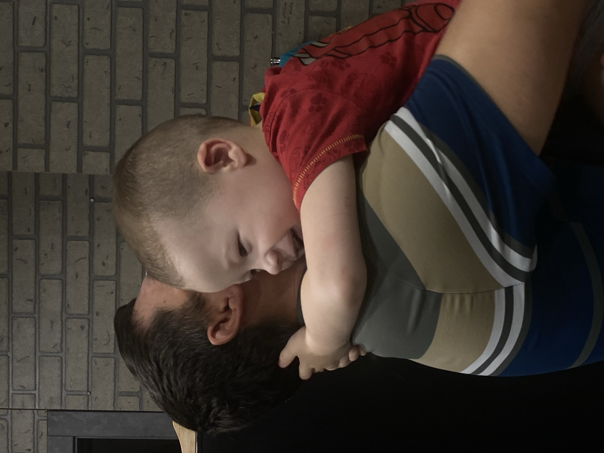 This image captures an indoor scene in Sochi, Russia, featuring an adult male holding a young child. The man, positioned with his back partially towards the camera, has dark brown hair and is wearing a short-sleeved, multi-striped garment in shades of olive green, white, gray, and blue. The child, appearing to be a toddler, is held by the man, with his back to the viewer, but his head is turned sharply to the left, revealing a partial profile. The child has light, short hair and is dressed in a red short-sleeved t-shirt with a faint embossed pattern and blue shorts or underwear. His arms are wrapped around the man's neck and right shoulder, suggesting an embrace or being carried. The child's expression displays an open-mouthed smile. The background consists of a dark gray brick wall with a faint, indecipherable inscription or marking on the right side and a small metallic hook. A dark, vertical object, possibly a door frame or furniture, is visible on the far left.
