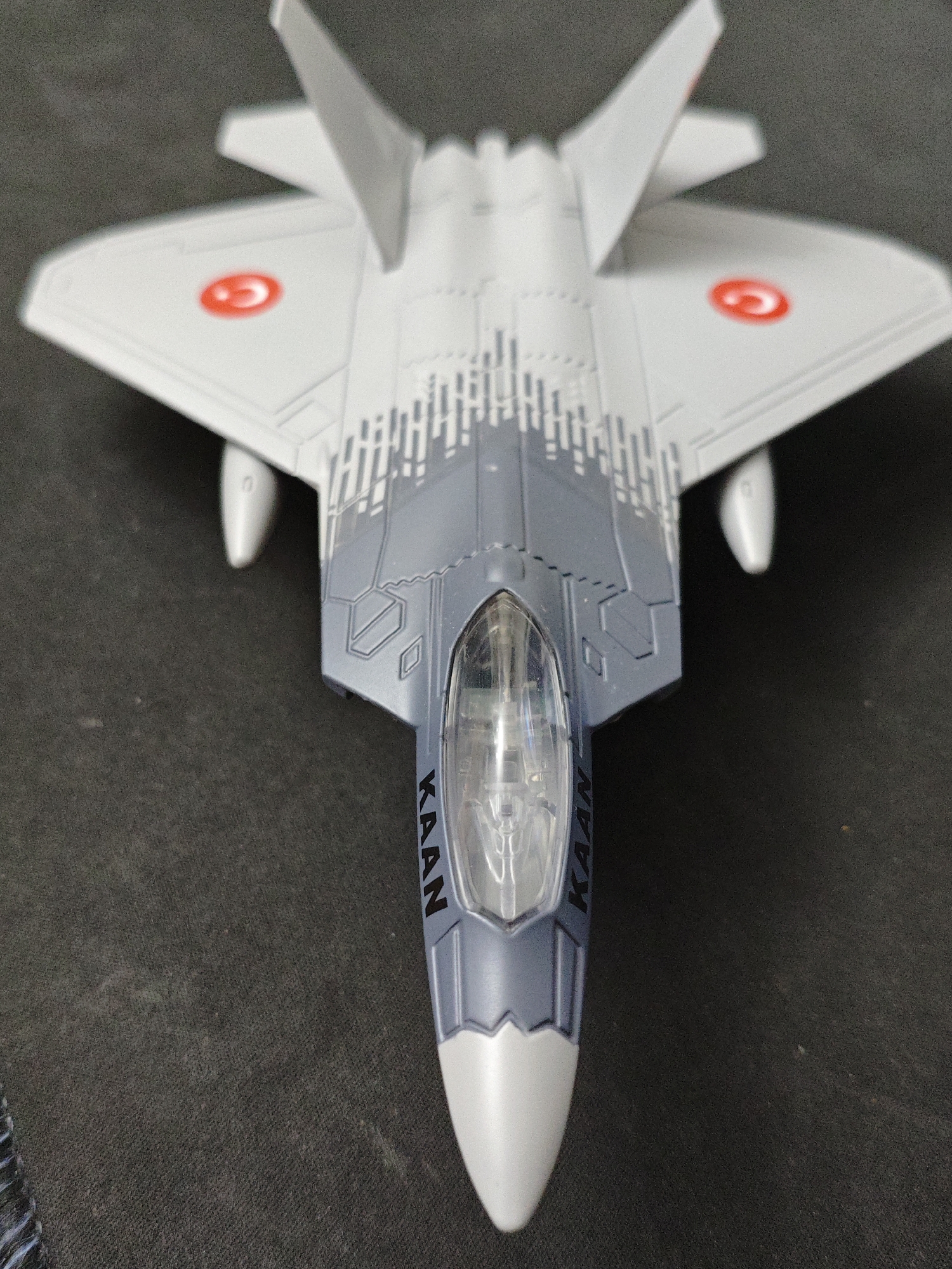 A top-down, slightly angled, close-up shot captures a model of a fighter jet, likely the Turkish Aerospace Industries TF-X "KAAN," displayed on a dark, textured surface, possibly a tablecloth or carpet. The model is predominantly white and gray, with intricate panel lines and detailing. Red circular emblems, resembling the Turkish flag, are visible on the wings. The cockpit canopy is clear, revealing internal detailing, and the word "KAAN" is printed vertically on the side of the fuselage near the canopy. The background is out of focus and appears to be a dark, uniform surface. The image does not contain any discernible elements that would indicate a specific location in Baku City, Azerbaijan.