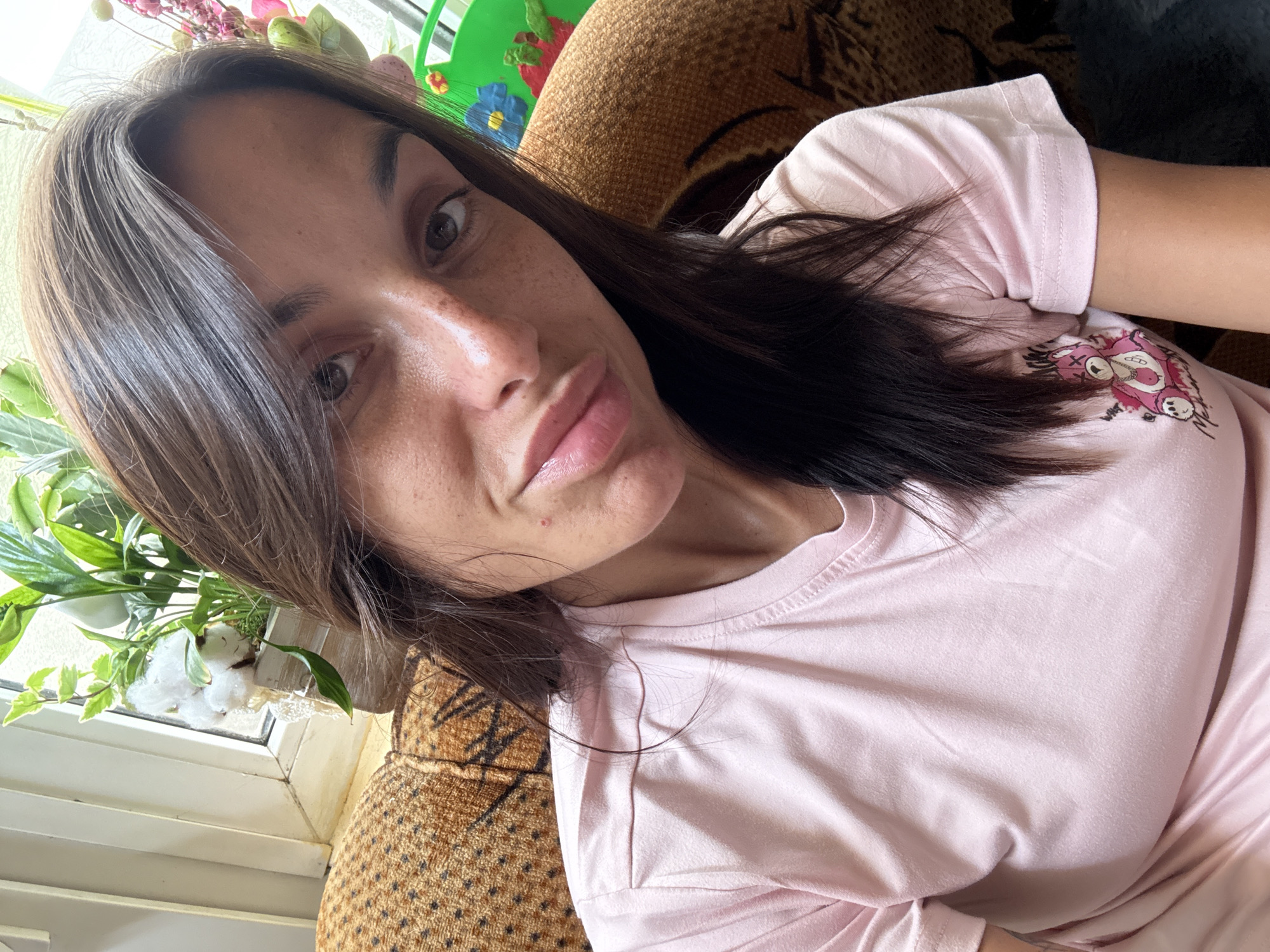 This is a close-up selfie of a woman with brown hair and brown eyes, wearing a pink t-shirt with a cartoon bear graphic. She has freckles on her face and a small pimple on her cheek. The background shows a patterned sofa, a window with plants, and a white curtain. The scene appears to be indoors, likely in a residential setting. The image does not provide any information about the city of Pleven, Bulgaria.