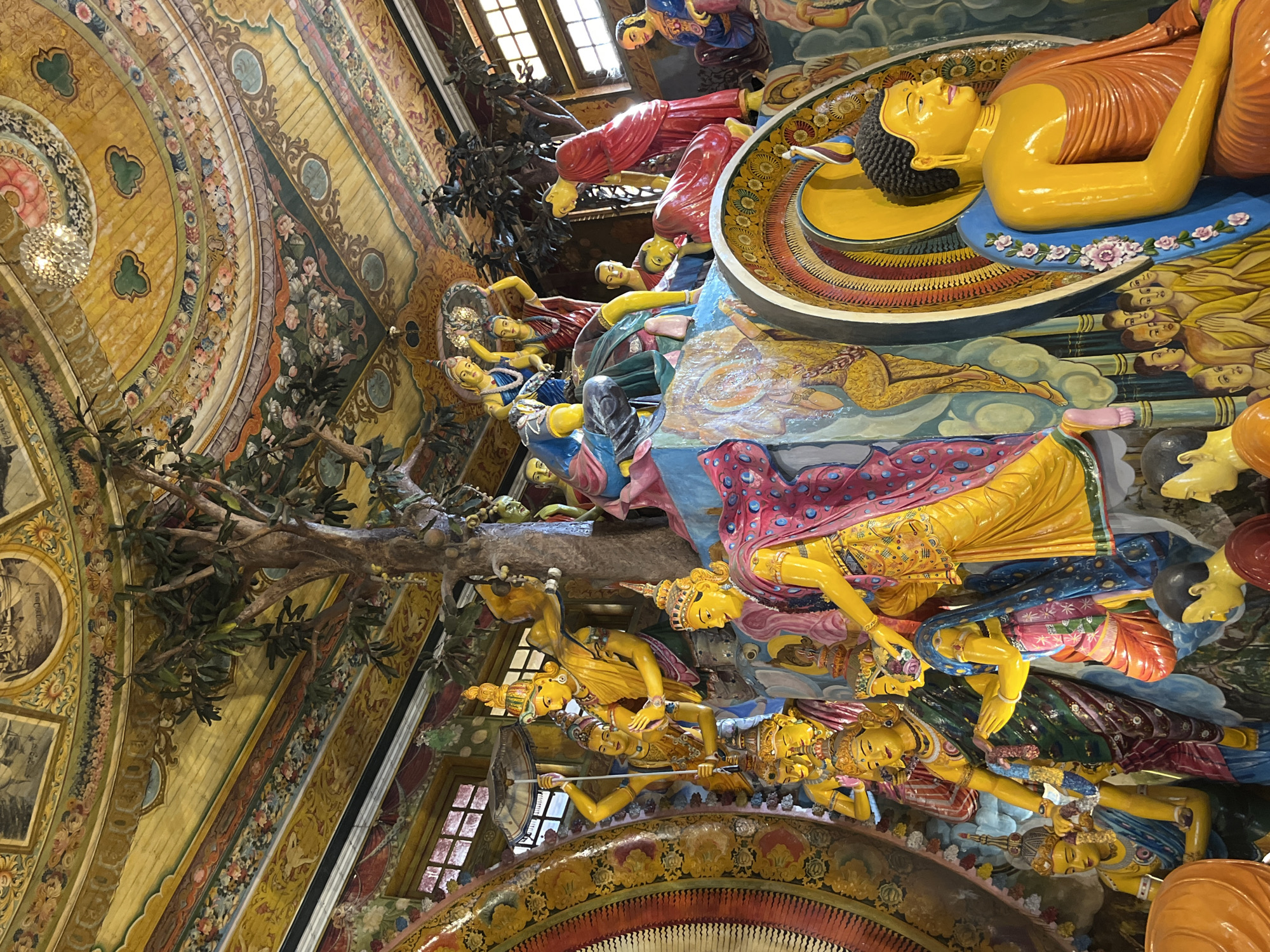 The image depicts a vibrant and ornate interior of a Buddhist temple in Colombo, Sri Lanka. The scene is dominated by colorful, elaborately decorated sculptures and murals, characteristic of Sri Lankan religious art. In the foreground, a large, golden Buddha statue sits with a serene expression, adorned in an orange robe. Behind and to the left of the Buddha, a complex arrangement of figures, likely depicting scenes from Buddhist mythology or history, unfolds. These figures are rendered in bright yellows, blues, reds, and greens, with many wearing crowns and flowing garments. A central, tree-like structure rises from the mid-ground, with sculpted figures interacting around its base. The ceiling is a marvel of intricate detail, painted with celestial motifs, floral patterns, and circular designs in shades of yellow, red, and blue, suggesting a highly sacred and revered space. The overall impression is one of rich spiritual and artistic heritage.