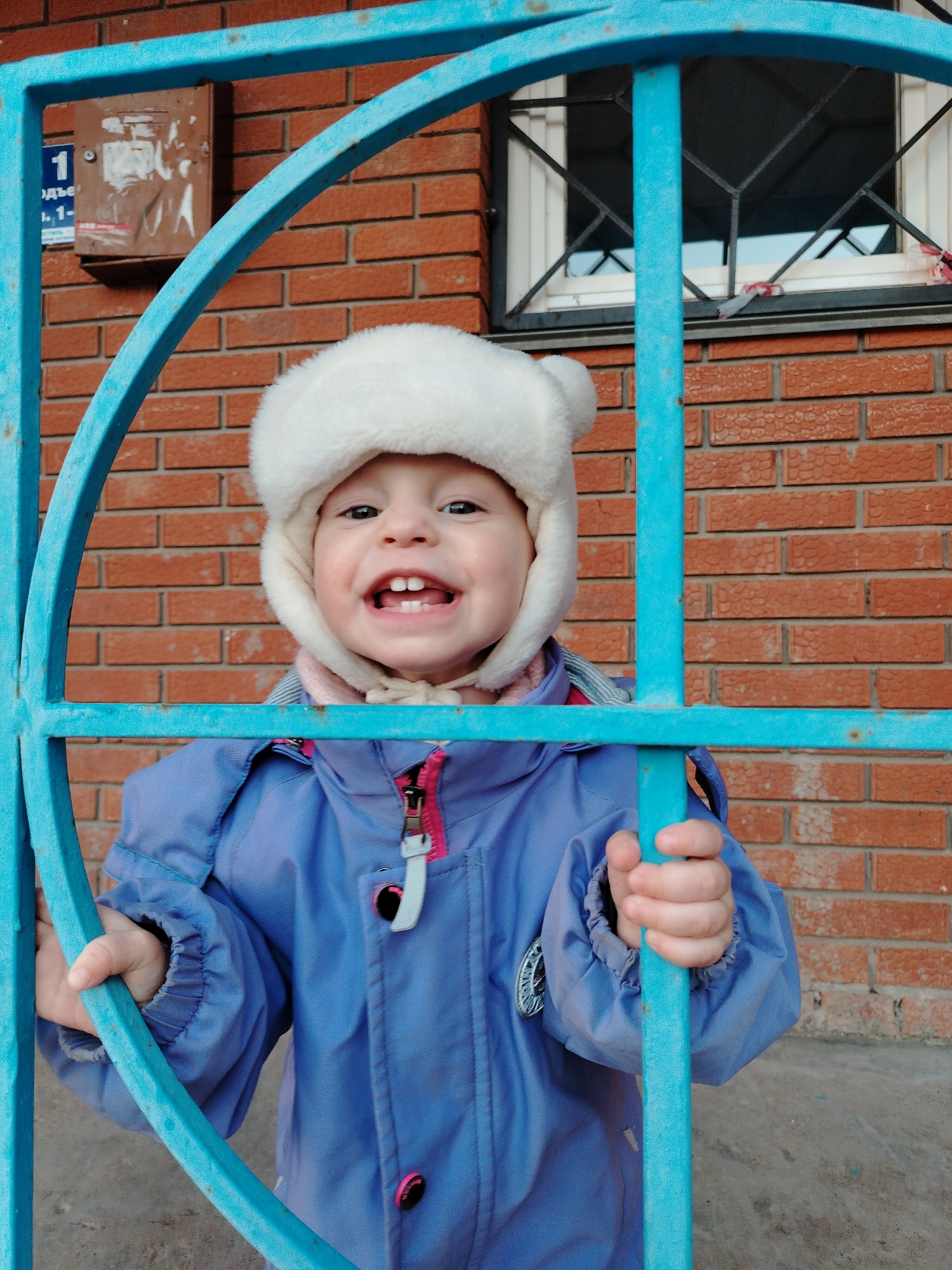 A child wearing a white winter hat with ear flaps and a blue jacket stands behind a blue metal gate in front of a brick building. The child is smiling and holding onto the bars of the gate. Behind the gate is a brick wall with a small window and a mailbox. The image was taken in Chaikovsky, Russia.