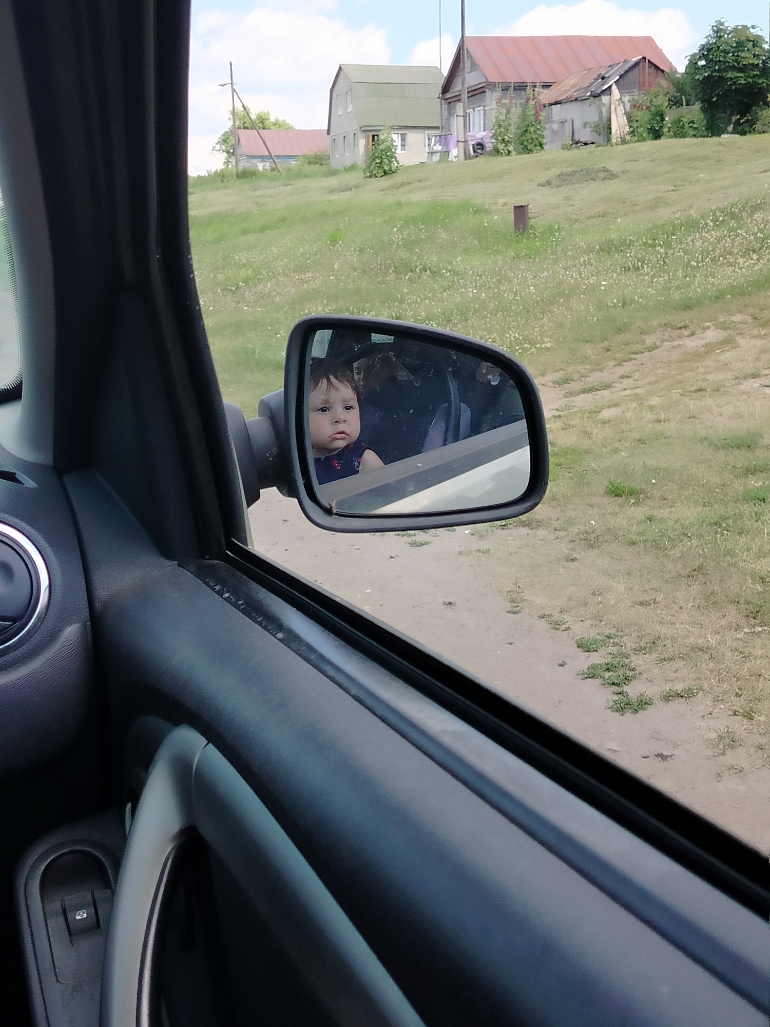 The image is taken from inside a vehicle, looking out of the passenger side window. A side-view mirror is prominent, reflecting a young child with a serious expression. The child appears to be wearing a dark blue garment. Outside the car, the scene depicts a rural landscape with a grassy hill dotted with wildflowers. In the background, several houses with red roofs are visible, suggesting a village or small town. A single utility pole stands on the left side of the frame. The lighting suggests it is daytime, and the weather appears to be overcast or partly cloudy. The overall impression is a snapshot of everyday life in a rural Russian setting, possibly during a car journey. No text is visible in the image.