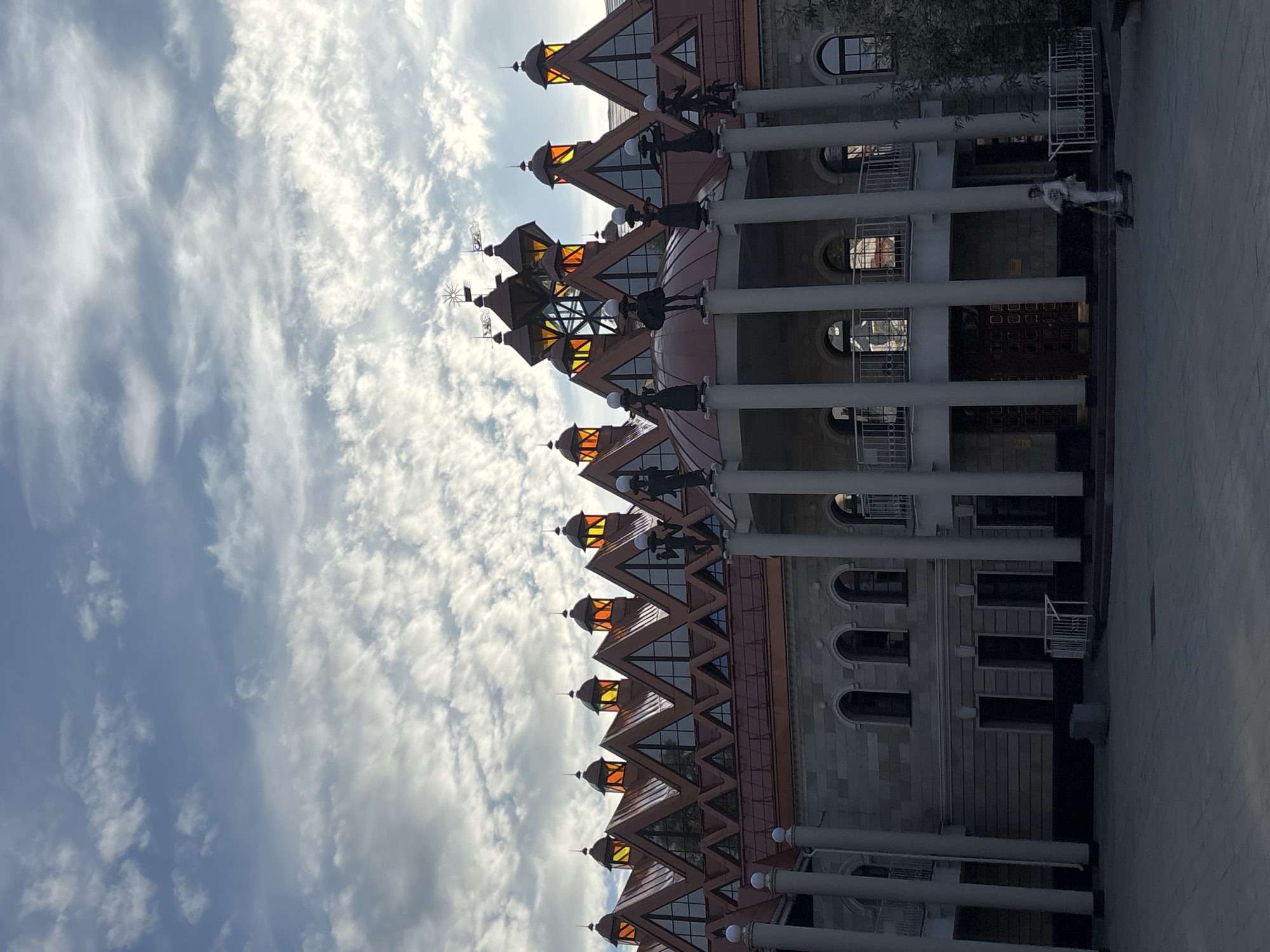 The image shows a striking building with a distinctive stepped roofline and triangular windows topped with orange illuminated panels. Sculptural figures stand atop pillars along the building's facade. A person is seen riding an electric scooter in the foreground on a paved plaza. The sky above is filled with dramatic, textured clouds. This building is identified as the Novaya Usman' Central District Library in Novaya Usman', Russia.