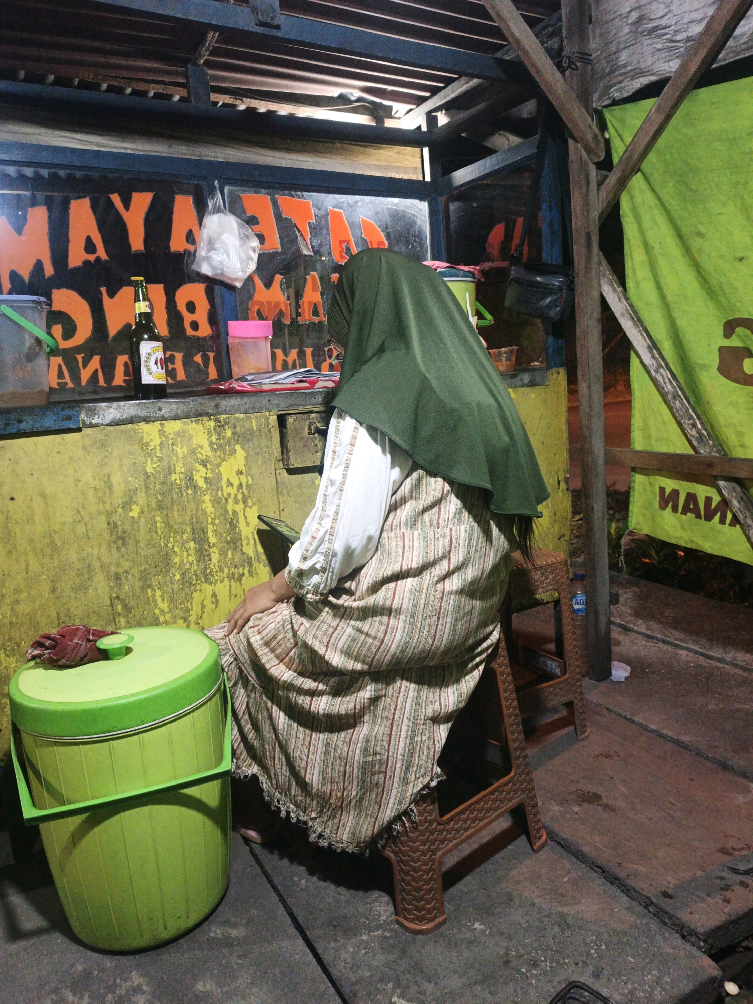 A person wearing a green hijab and a patterned skirt sits on a brown plastic stool. The person is facing away from the camera and appears to be holding a cell phone. To the left of the person is a large green plastic cooler with a red cloth draped over the lid. Behind the person is a yellow wall with orange lettering. A bottle of beer and some plastic containers are on a counter in front of the wall. A green tarp hangs on a wooden structure to the right of the scene. The scene appears to be an outdoor food stall or a similar roadside establishment in Balongmojo, Indonesia. The ground is concrete and appears to be worn.