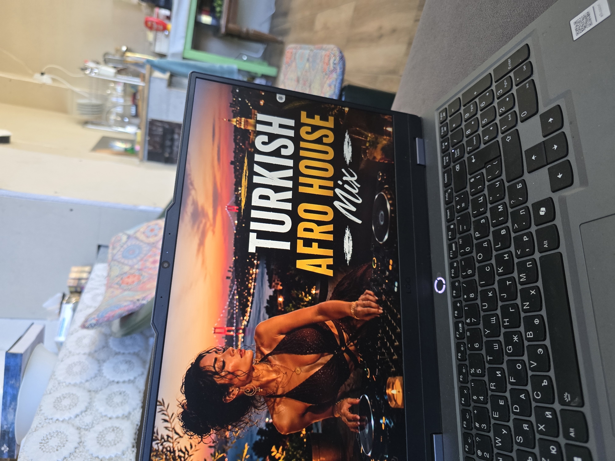 The image shows a laptop on what appears to be a grey couch. The laptop screen displays an image of a woman in a dress in front of a city skyline with the text "TURKISH AFRO HOUSE Mix". The laptop has a grey keyboard with Cyrillic characters in addition to the standard alphabet. The background shows part of a room including a table with items on it, a wall and chair. There is nothing to indicate this photo was taken in Dorkovo, Bulgaria.
