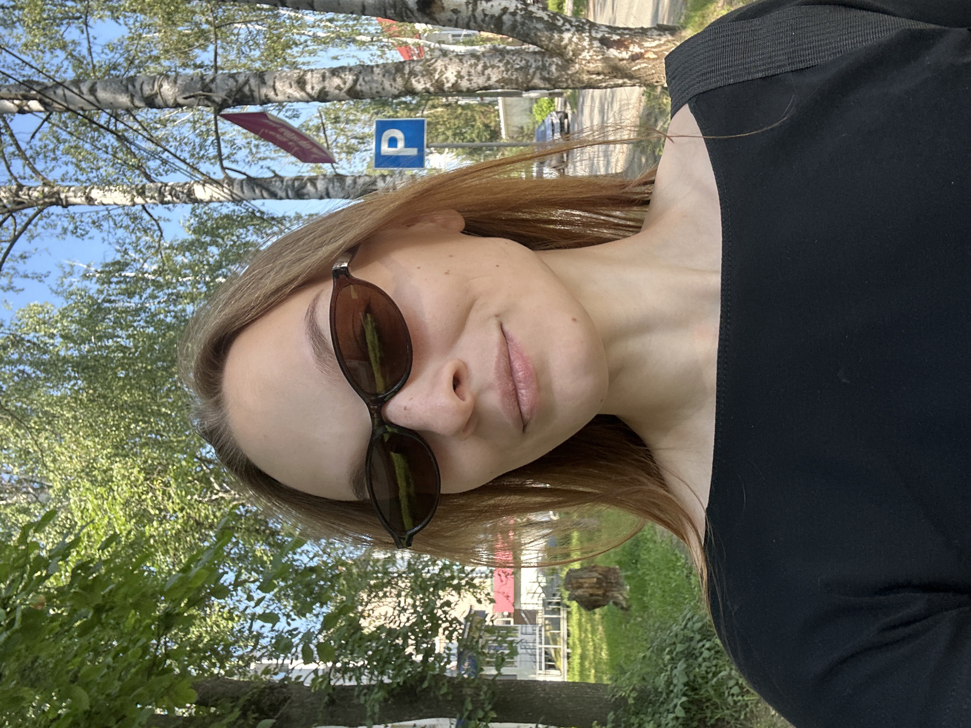 A woman is taking a selfie outdoors in Chaikovsky, Russia. She is wearing sunglasses and a black shirt, and her hair is light brown. In the background, there are trees, buildings, and a blue parking sign. The weather appears to be sunny.