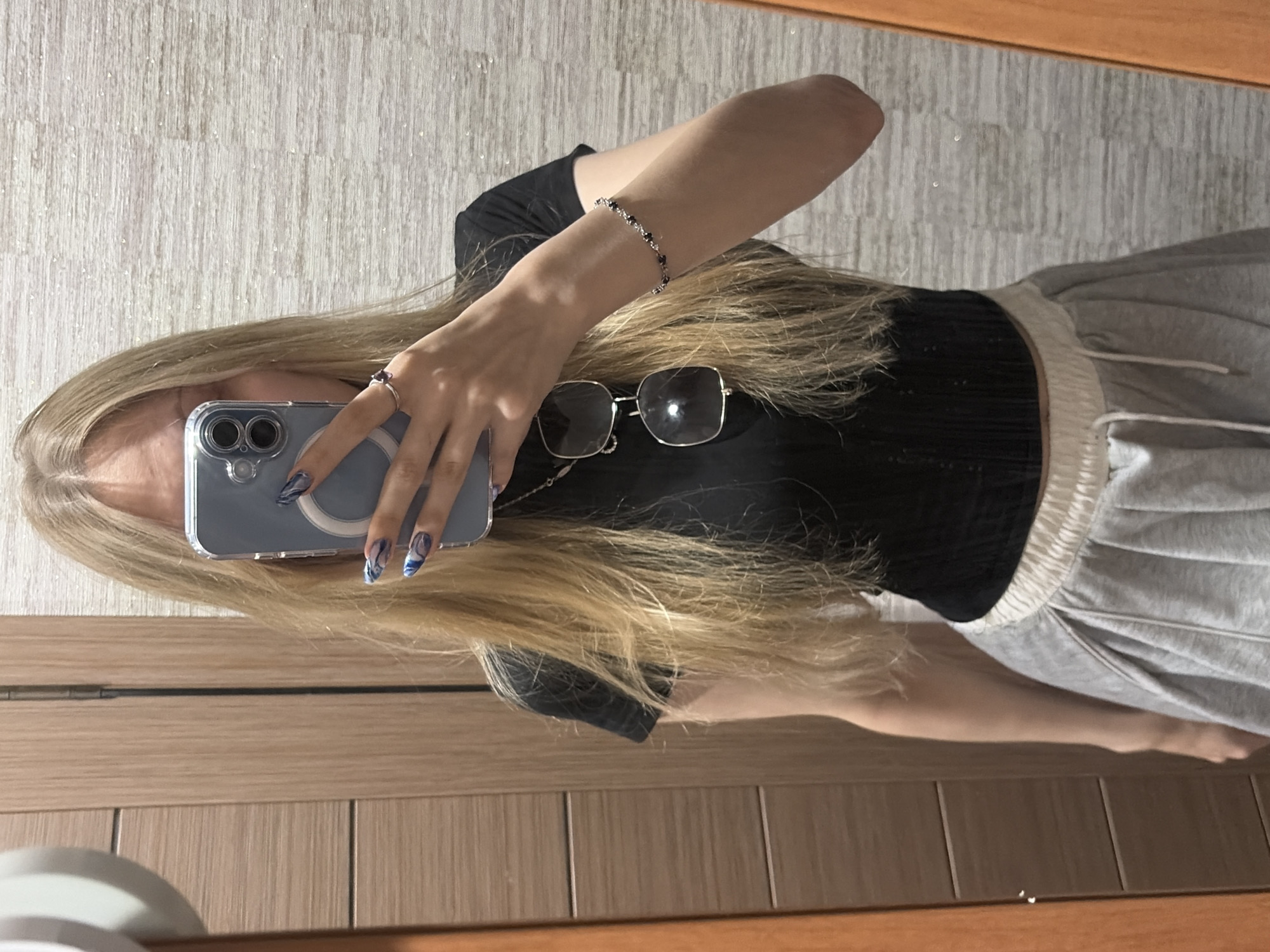 A person stands in front of a mirror, holding a phone to take a selfie. The person has long, blonde hair and is wearing a black top and a gray skirt. They are also wearing a ring on their right hand and a bracelet on their left wrist. Sunglasses are hanging from their neck. The background appears to be a room with wooden doors and patterned wallpaper. The overall scene is casual and domestic.