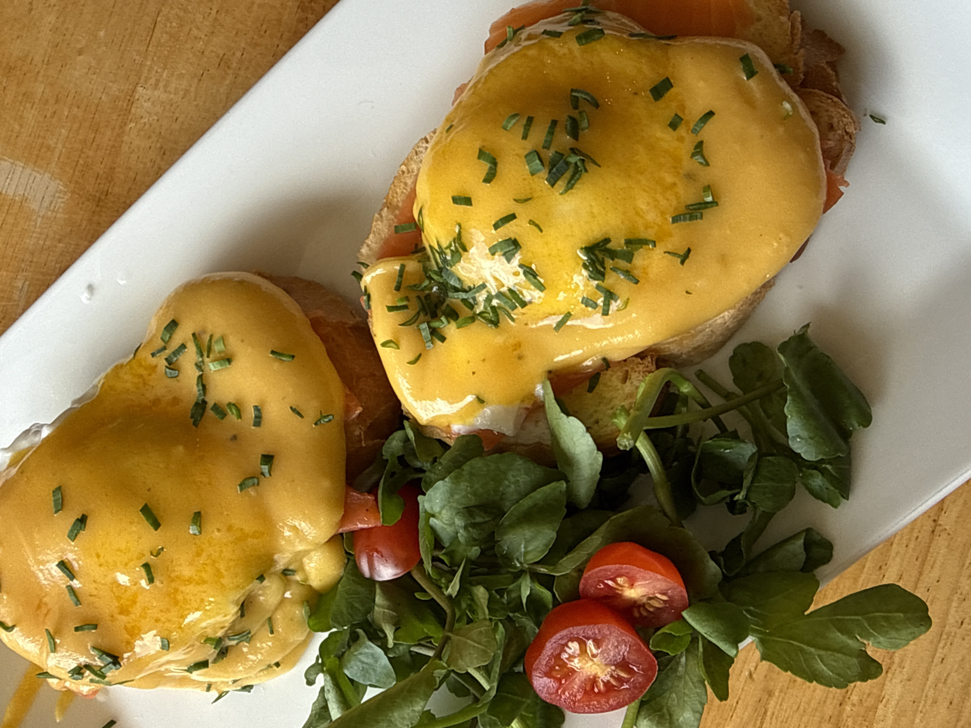 Two dishes of Eggs Benedict are presented on a white rectangular plate, which rests on a light brown wooden surface. Each dish features a toasted bread base topped with smoked salmon, a poached egg, and hollandaise sauce. Chopped chives are sprinkled over the hollandaise sauce for garnish. To the left of the dishes, a small portion of a green salad with halved cherry tomatoes is visible. The image is a close-up, overhead shot, with the focal point on the food. There are no people or animals in the image, and the environment suggests an indoor setting, likely a restaurant. The specific location in Da Lat, Vietnam cannot be determined from the visual information provided.