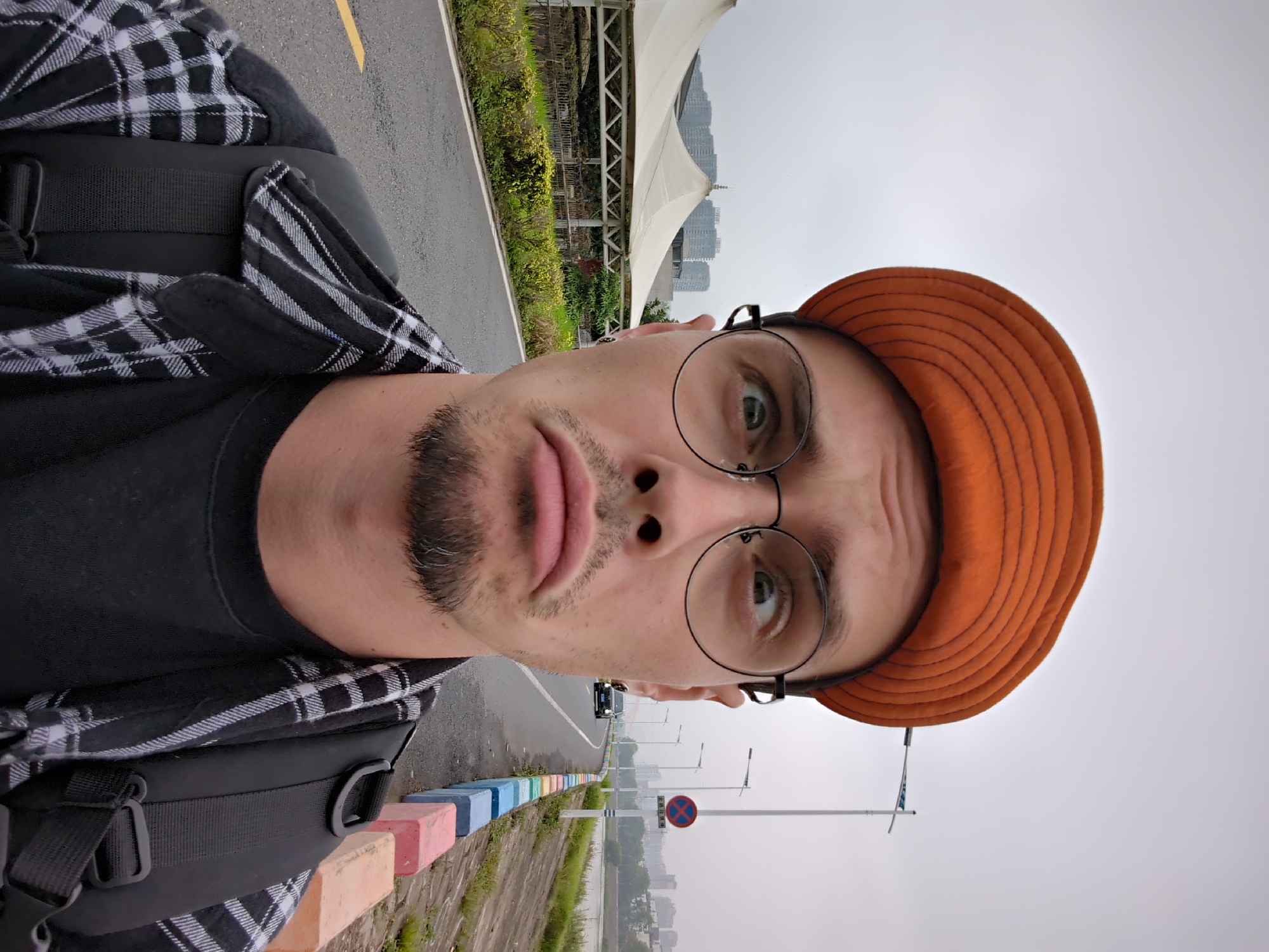 A man is taking a selfie. He has light skin, a beard, and is wearing round glasses and an orange corduroy cap. He has on a black t-shirt and a black and white plaid shirt over it. He is wearing a black backpack.
He is standing on a concrete path next to a road with multicolored concrete blocks separating the path from the road. There is a no stopping sign on a pole. There is a body of water to the side and buildings in the distance.
This is the city of Shunde District, China.