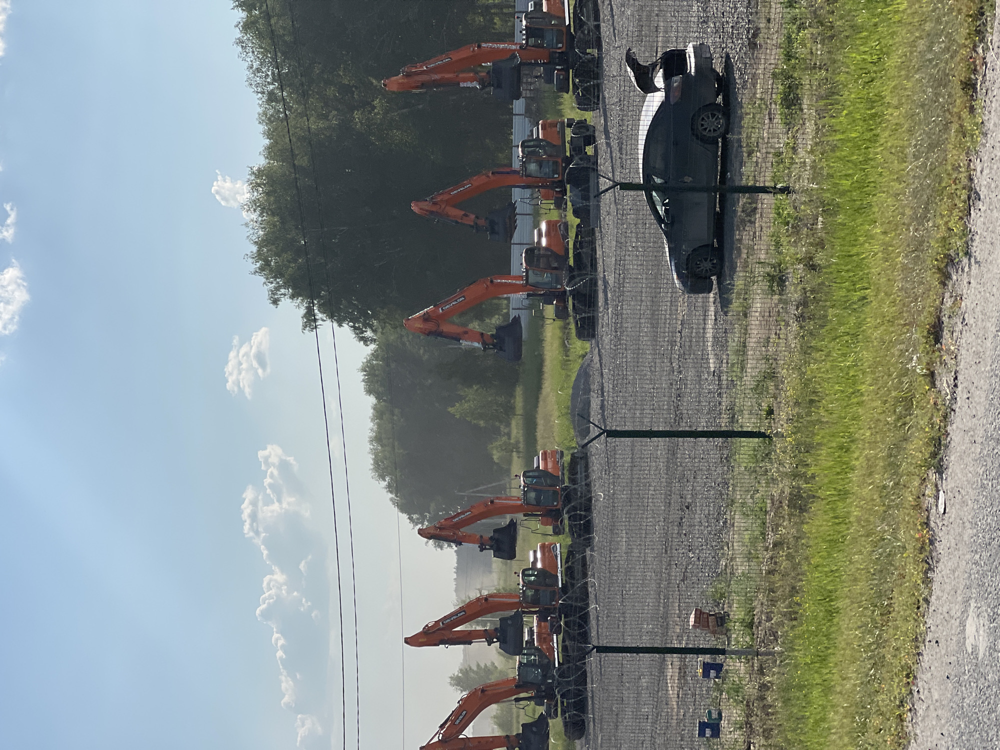 The image shows a row of orange excavators parked on a gravel lot. A dark-colored sedan is parked to the right of the excavators, with its driver's side door open and a person's arm visible reaching into the car. The scene is outdoors, with a fence separating the lot from a grassy area. In the background, there are trees and a bright blue sky with scattered white clouds, suggesting it is daytime. The lighting indicates it might be late morning or early afternoon. The excavators appear to be new or well-maintained, all facing in the same direction. The presence of multiple excavators suggests a construction site, equipment rental yard, or similar operational area. The open car door and the arm reaching in imply someone is either getting in or out of the vehicle, or retrieving something from it. No specific text is clearly legible on the excavators or the car. The overall impression is one of a functional, operational outdoor space.
