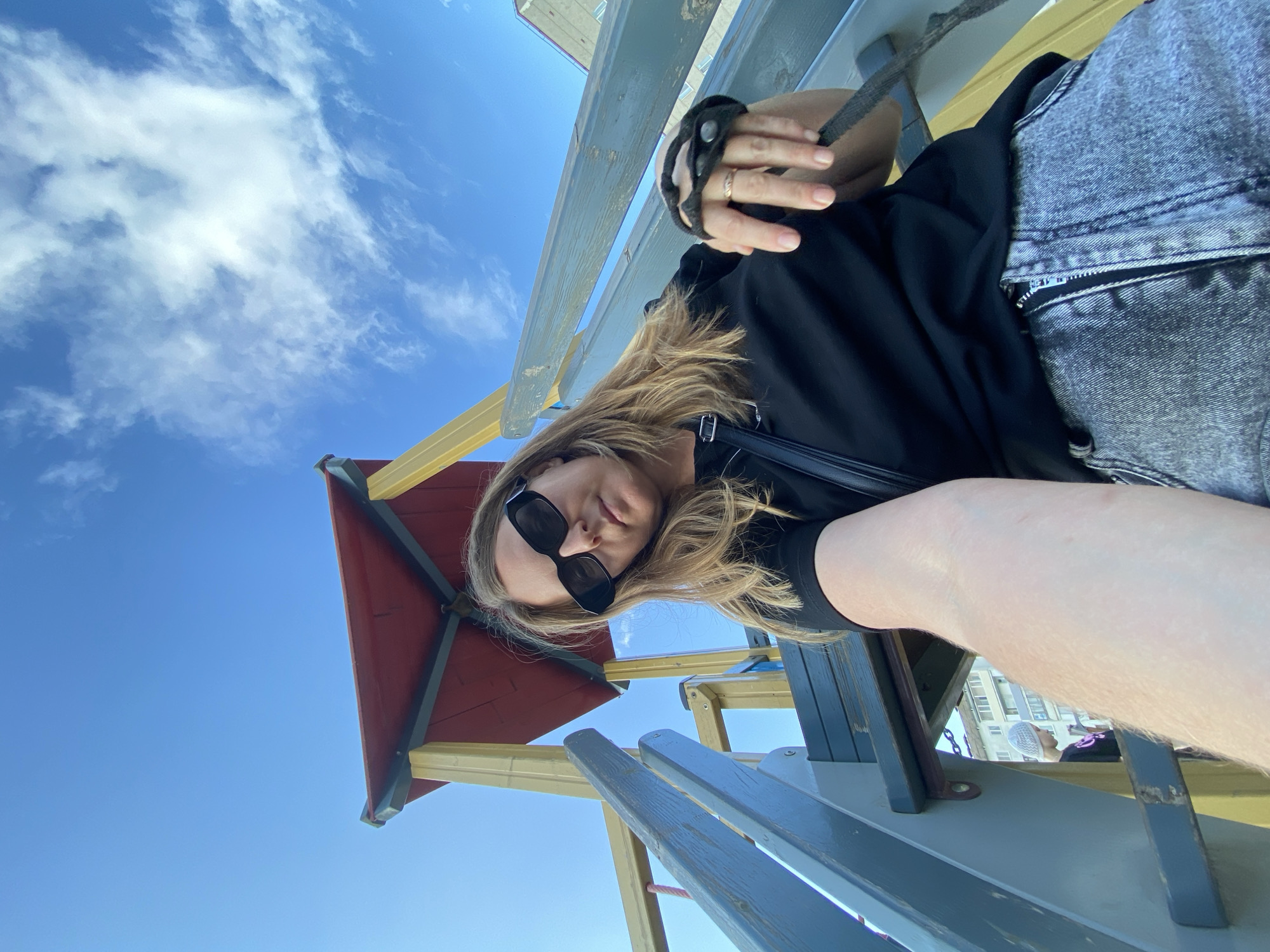 In Noviy, Russia, a woman with blonde hair and sunglasses takes a selfie on a playground. She is wearing a black t-shirt and grey jeans. A red and yellow playground structure is visible behind her. The sky is blue with some white clouds. A child in a light-colored hat is partially visible in the background.