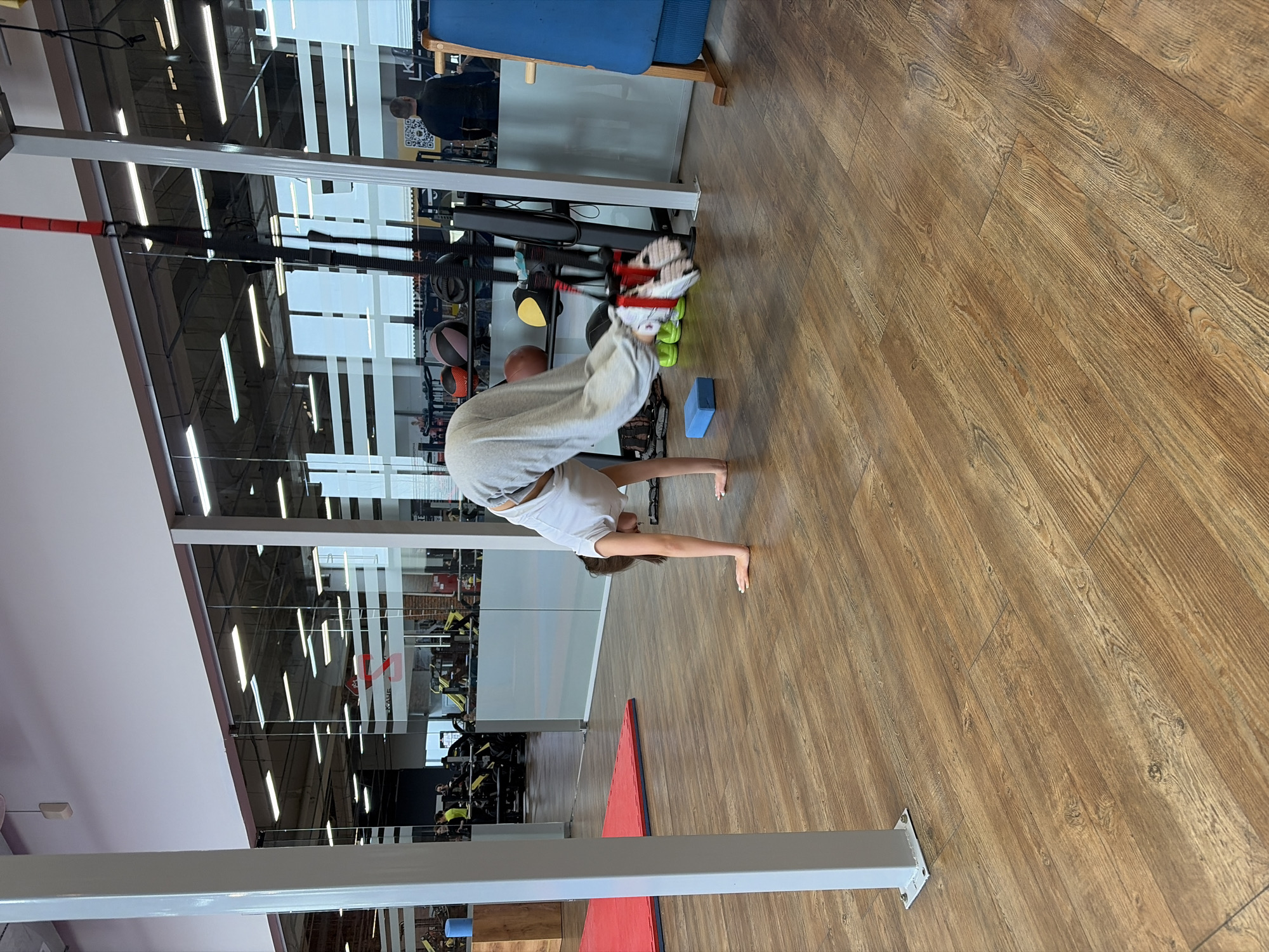 A person is performing a headstand in what appears to be a gym or fitness center. The person is wearing light-colored shorts and a grey sweatshirt. Their hands are on the floor, supporting their body. The fitness center has wood-look flooring and glass walls. Exercise equipment is visible in the background. The location is Chaikovsky, Russia.