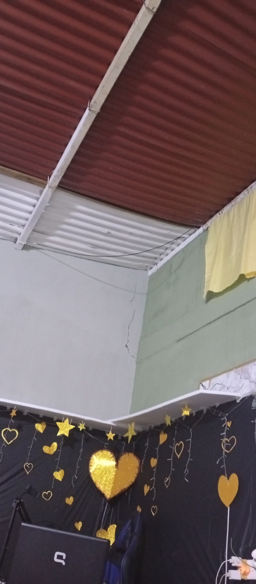 The image depicts an indoor scene, seemingly in a room, potentially in Juan Griego, Venezuela. The ceiling is comprised of two sections: one with a corrugated red-brown metal roof and the other with white corrugated metal sheets supported by white beams. A crack is visible on the pale green wall. Below the wall, a black curtain-like backdrop is adorned with gold stars and heart decorations. The curtain seems to be set up for an event. A laptop with a white logo is partially visible on what seems to be a table in front of the backdrop. A yellow cloth is hanging in the upper right-hand corner of the frame.