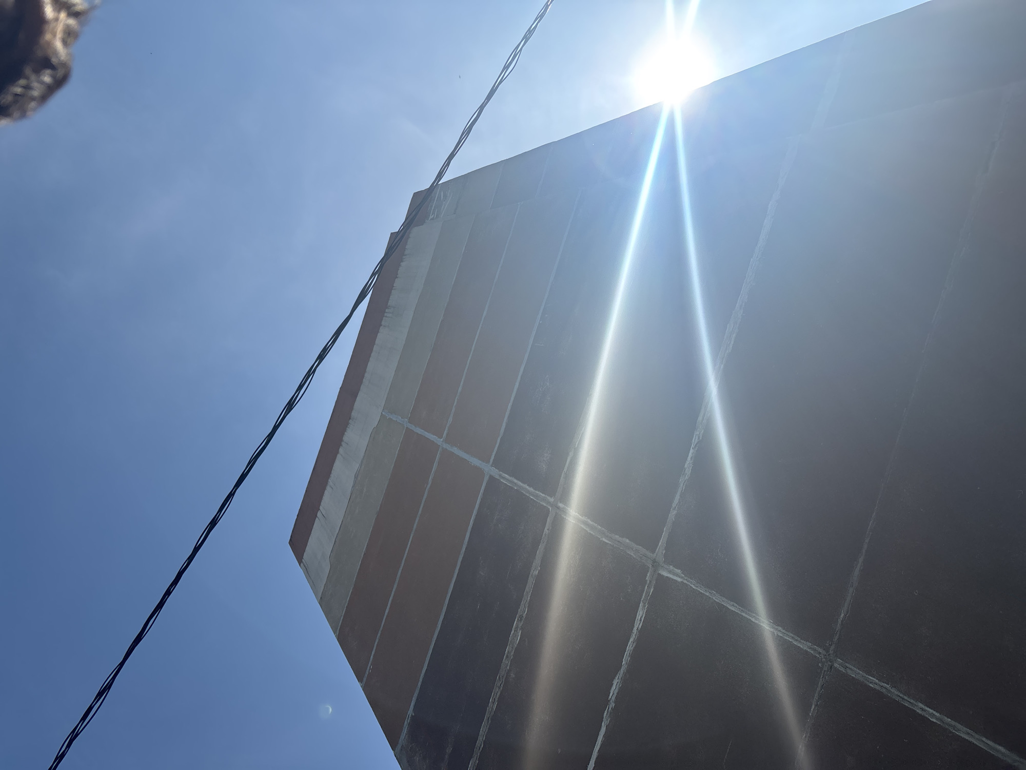The image captures a sunlit building in Minsk, Belarus, with a clear blue sky. The building's facade is segmented into rectangular panels, with varying shades of brown and gray, suggesting potential aging or different materials. Power lines are visible crisscrossing the sky in the foreground, with one set extending diagonally across the top of the frame and another further down and to the right. The sun is intensely bright, positioned in the lower right quadrant of the image, causing significant lens flare and glare that obscures parts of the building and sky. A sliver of the moon is faintly visible in the upper left quadrant against the blue sky. At the very top right edge of the frame, a blurry, dark object, possibly part of another structure or an aerial element, is partially visible. The perspective is from below, looking up at the building.