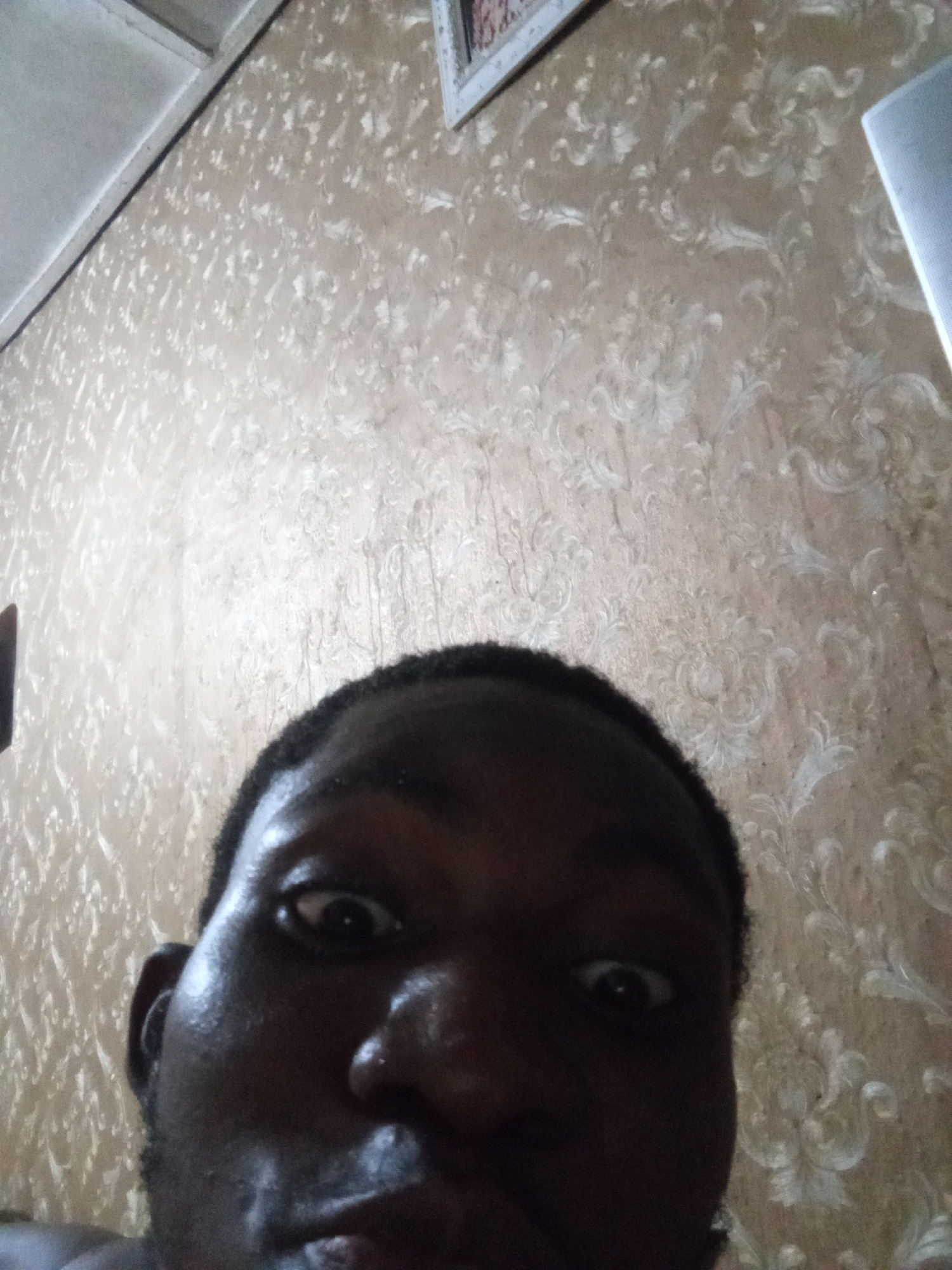 A close-up selfie of a dark-skinned person with short, dark hair. The person's face is the primary focus, with eyes wide and a slightly surprised expression. The background is a wall with a floral pattern wallpaper. There are visible elements of a ceiling and frame. The location is unknown.