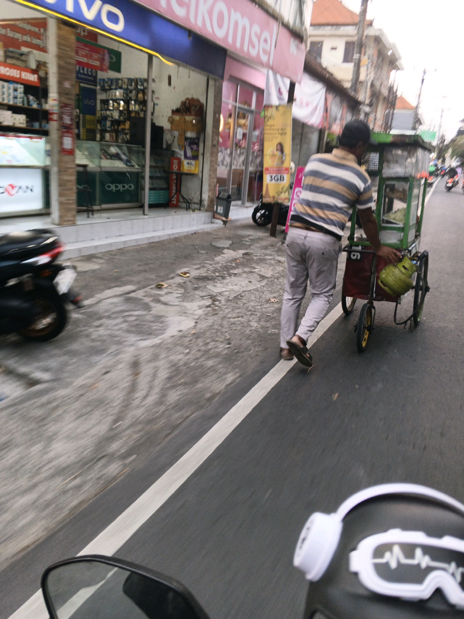 In Denpasar, Indonesia, a man pushes a street food cart along a paved road. He wears a horizontally striped shirt and light-colored pants, with sandals on his feet. The cart is green and red, equipped with a yellow propane tank and various items for sale, including what appears to be fresh produce. A black motorcycle is parked on the left side of the street in front of a shop. The shop displays "VIVO" and "OPPO" branding, suggesting it is an electronics store. The overall scene depicts a typical street environment in Denpasar, with shops, vehicles, and street vendors.