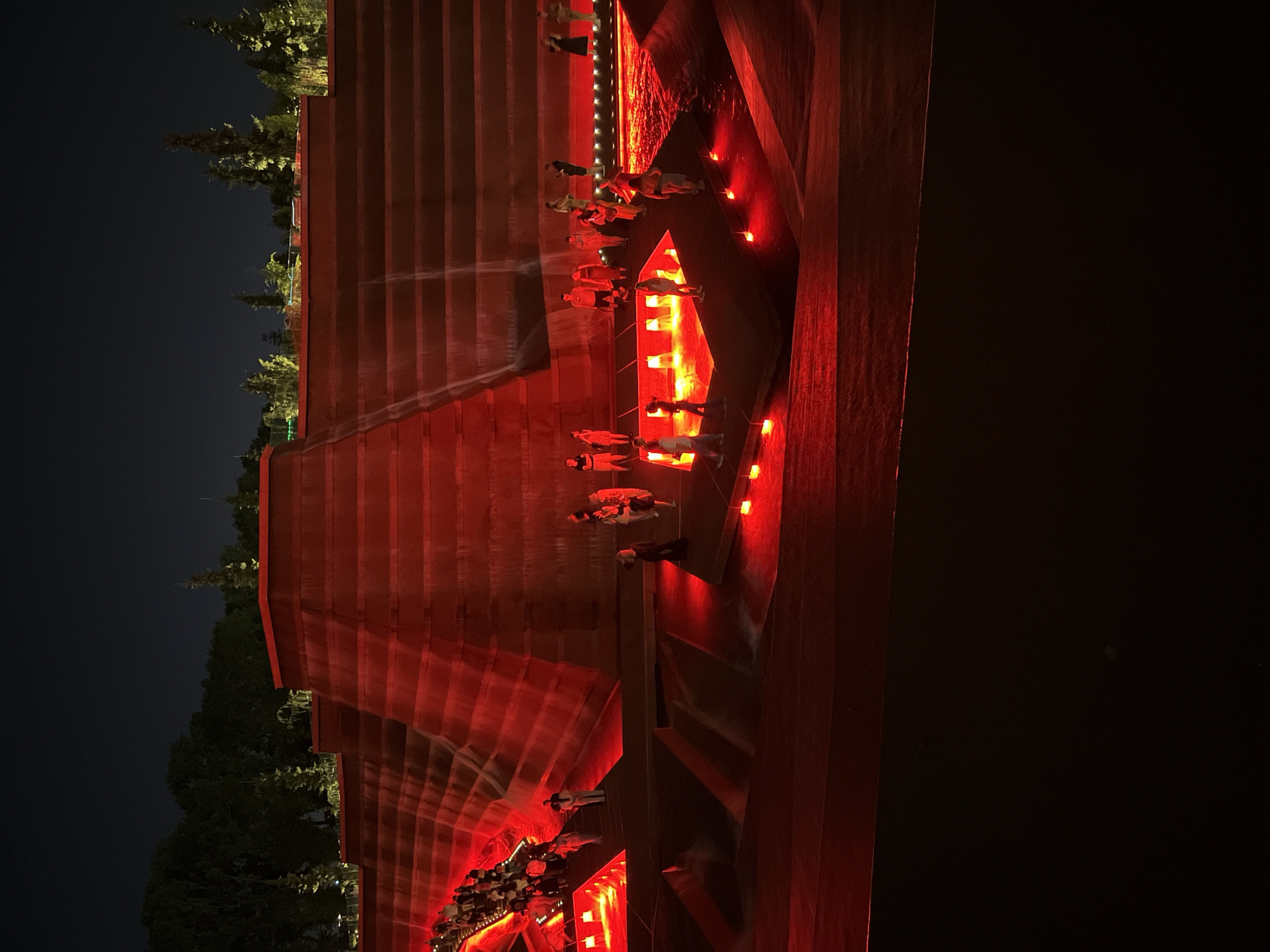 The image shows a night scene at the "Waterfront" park in Krasnodar, Russia. The park features a multi-tiered, red-lit waterfall structure with people gathered at various levels. Several individuals are visible standing on the illuminated tiers and near the water features, some appearing to be observing the spectacle. The lighting is predominantly red, creating a dramatic and artificial atmosphere. Trees are visible in the background, silhouetted against the dark night sky. The water cascades down the red-lit tiers, reflecting the light.