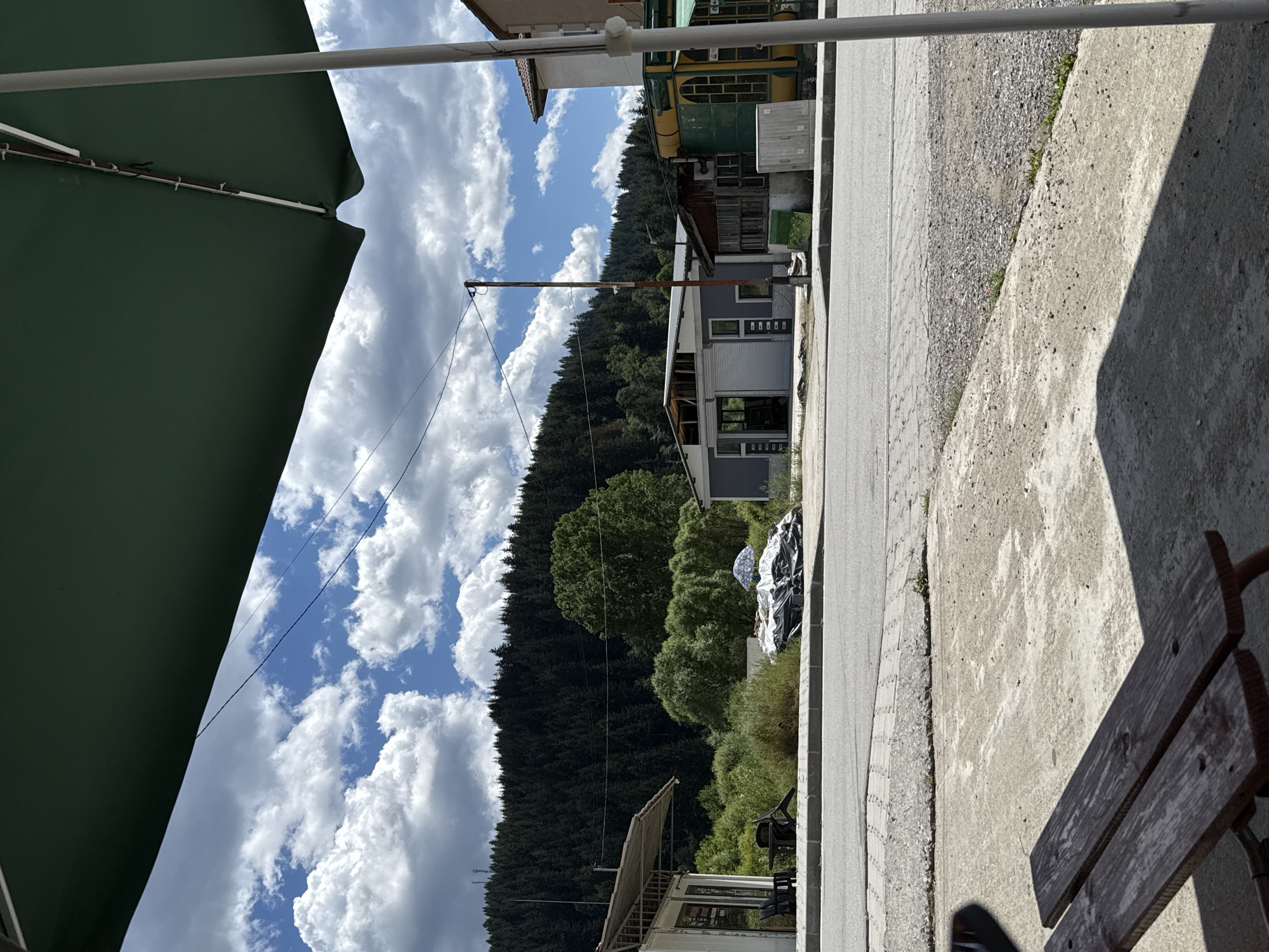 In the city of Barutin, Bulgaria, the image captures a roadside scene under a large, dark green umbrella. In the foreground, a wooden bench is partially visible, casting a shadow on a concrete surface. Across a paved road, several buildings line the street. To the left, a small white structure with an awning stands, with two chairs and a table outside. Further down the street, a grey building with an open roof section is visible, alongside other structures featuring green and yellow accents. Behind the buildings, a dense forest covers a hillside that rises to meet a cloudy sky. The environment appears to be a quiet, rural area.