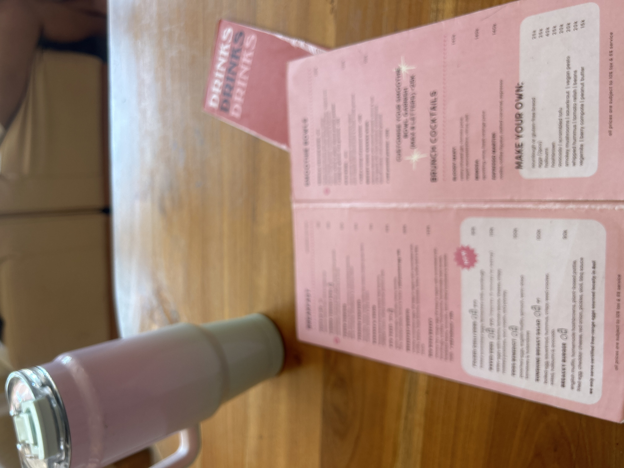 The image shows a pink menu on a wooden table, with a pink and white insulated tumbler in the background. The menu is divided into sections with text in Thai and English, including "BREAKFAST," "SMOOTHIE BOWLS," "BRUNCH COCKTAILS," and "MAKE YOUR OWN." The tumbler has a handle and a clear lid with a light-colored spout. A red card with the word "DRINKS" repeated in white font is placed behind the menu. The setting appears to be an indoor cafe or restaurant, likely in Kuta Selatan, Indonesia, as suggested by the context. There are no people visible in the image, and the focus is on the menu and the tumbler.