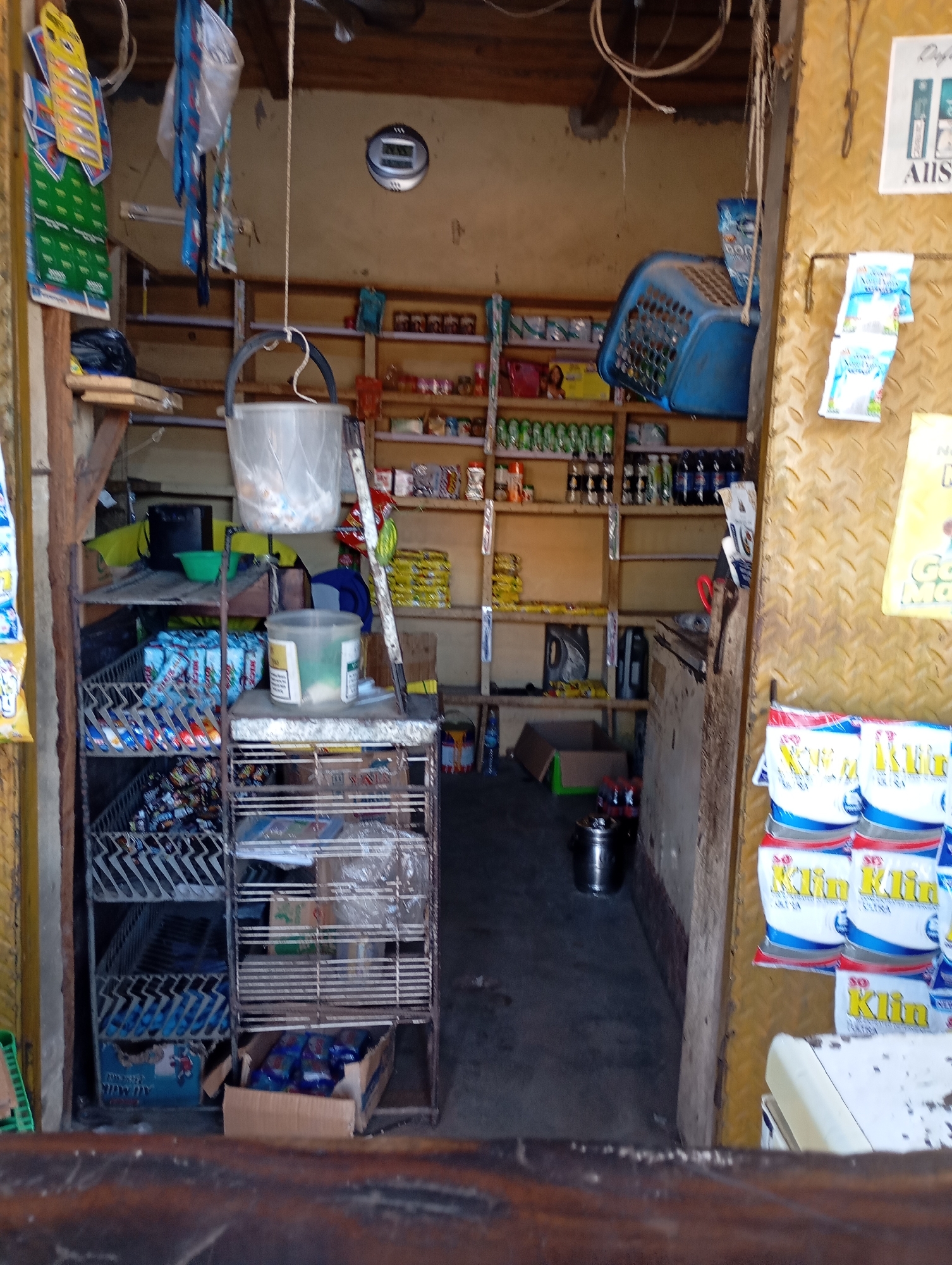 The image depicts the interior of a small shop, likely a convenience store or kiosk, located in Shendam, Nigeria. The shop is filled with various products displayed on shelves and racks. To the left, there are hanging items which appear to be small packaged goods or promotional materials. A large clear plastic bucket is suspended from the ceiling. Shelves are stacked with a variety of items, including what look like bottled drinks, packaged snacks, and household products. A metal shelving unit is prominent, holding numerous packaged items, possibly snacks or detergent packets. In the foreground, a wooden counter or ledge is visible. The walls appear to be unfinished, with exposed wood and concrete. The overall impression is of a modestly stocked, functional retail space.