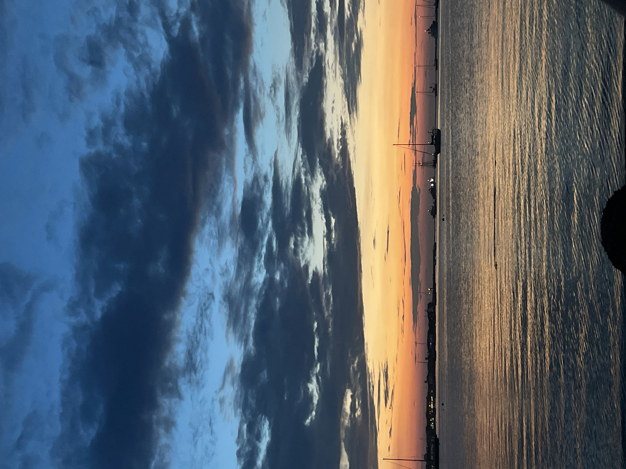 The image depicts a serene coastal scene on the island of Nantucket, USA, during twilight. The sky is a dramatic canvas of deep blues and grays at the top, transitioning to vibrant oranges and yellows near the horizon, characteristic of a sunset. Scattered clouds, tinged with dark hues, add texture to the sky.
The foreground is dominated by a calm body of water, likely the harbor or bay, with gentle ripples reflecting the colors of the sky. In the mid-ground, a line of sailboats and larger vessels are anchored, their masts and structures silhouetted against the fading light. Some of these boats have their navigation lights illuminated, adding small points of light to the scene. Along the distant shoreline, a town is visible, with buildings and lights suggesting human habitation. The overall impression is one of tranquility and the peaceful end of a day. There are no visible people in the frame. The environment is natural and maritime, with the water and sky as the primary elements.