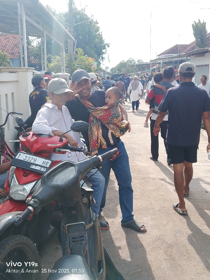 A casual street scene depicts a man carrying a baby secured in a fabric sling, flashing a peace sign for the camera. An older man sits on a red scooter beside him. The image features a black scooter parked in front and partially obstructing the view. A crowd of people strolls in the background of a sunlit street lined with houses. The bottom corner displays text: "vivo Y19s Akhtar & Arvan 25 Nov 2025, 07.53".